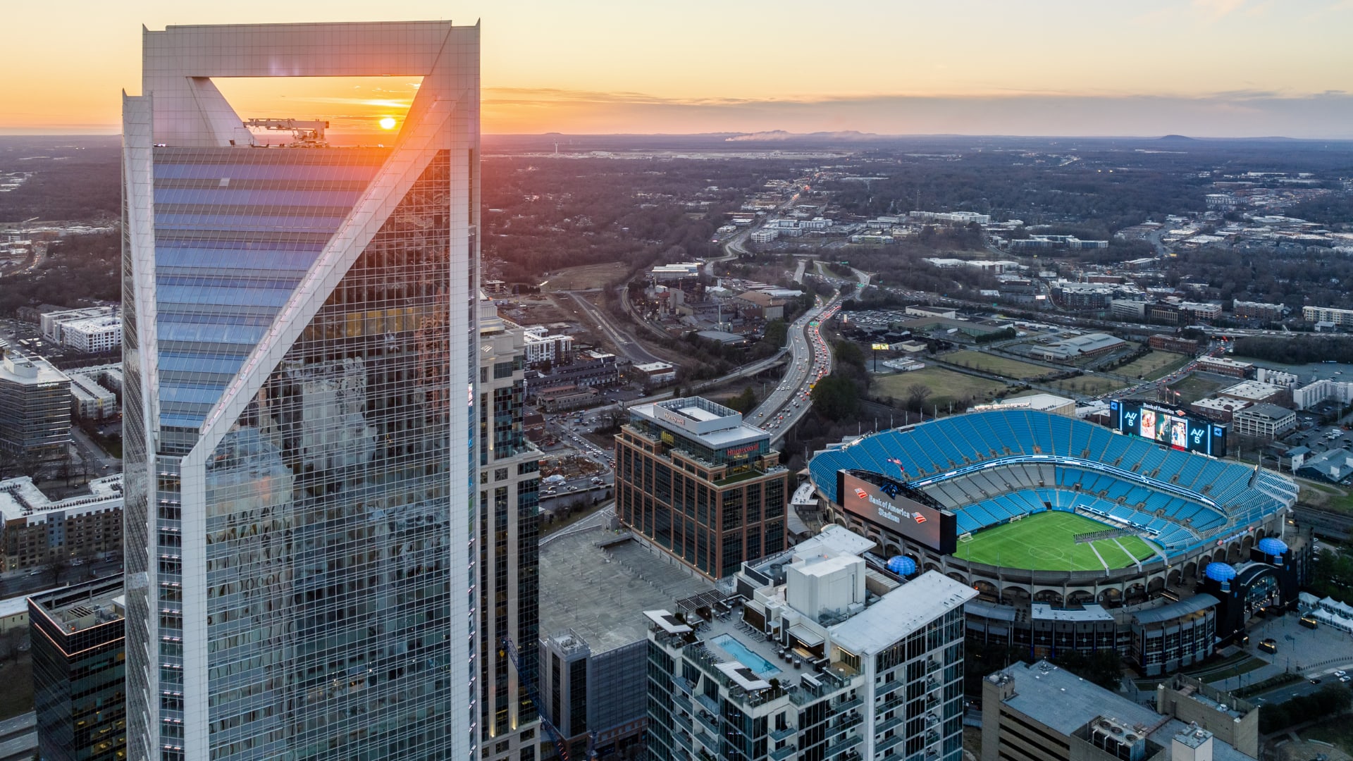 A modern city skyline with a large glass-fronted skyscraper in the foreground, reflecting the setting sun, and a sports stadium visible in the background against a mountainous landscape.