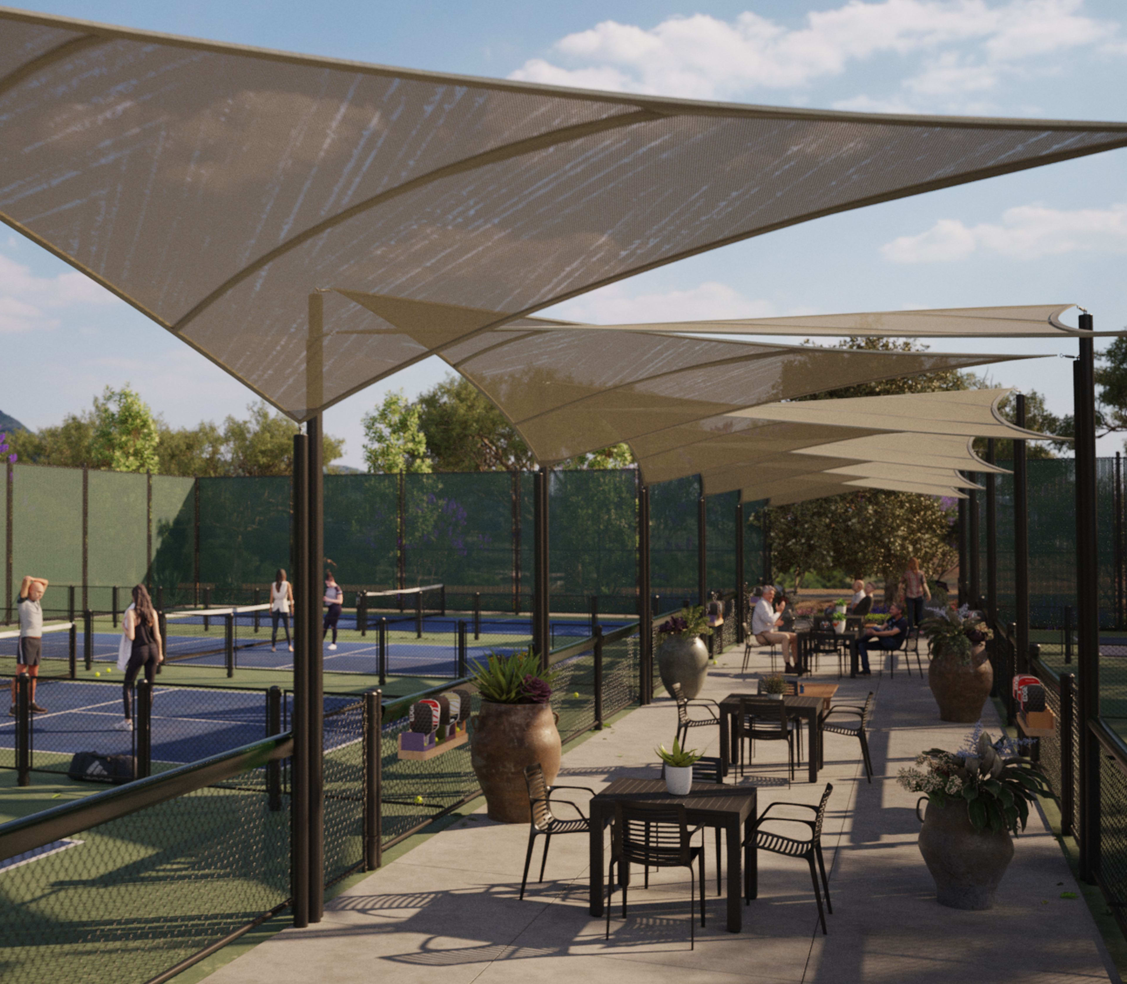 A shaded outdoor seating area with tennis courts in the background, surrounded by lush greenery and a clear blue sky.