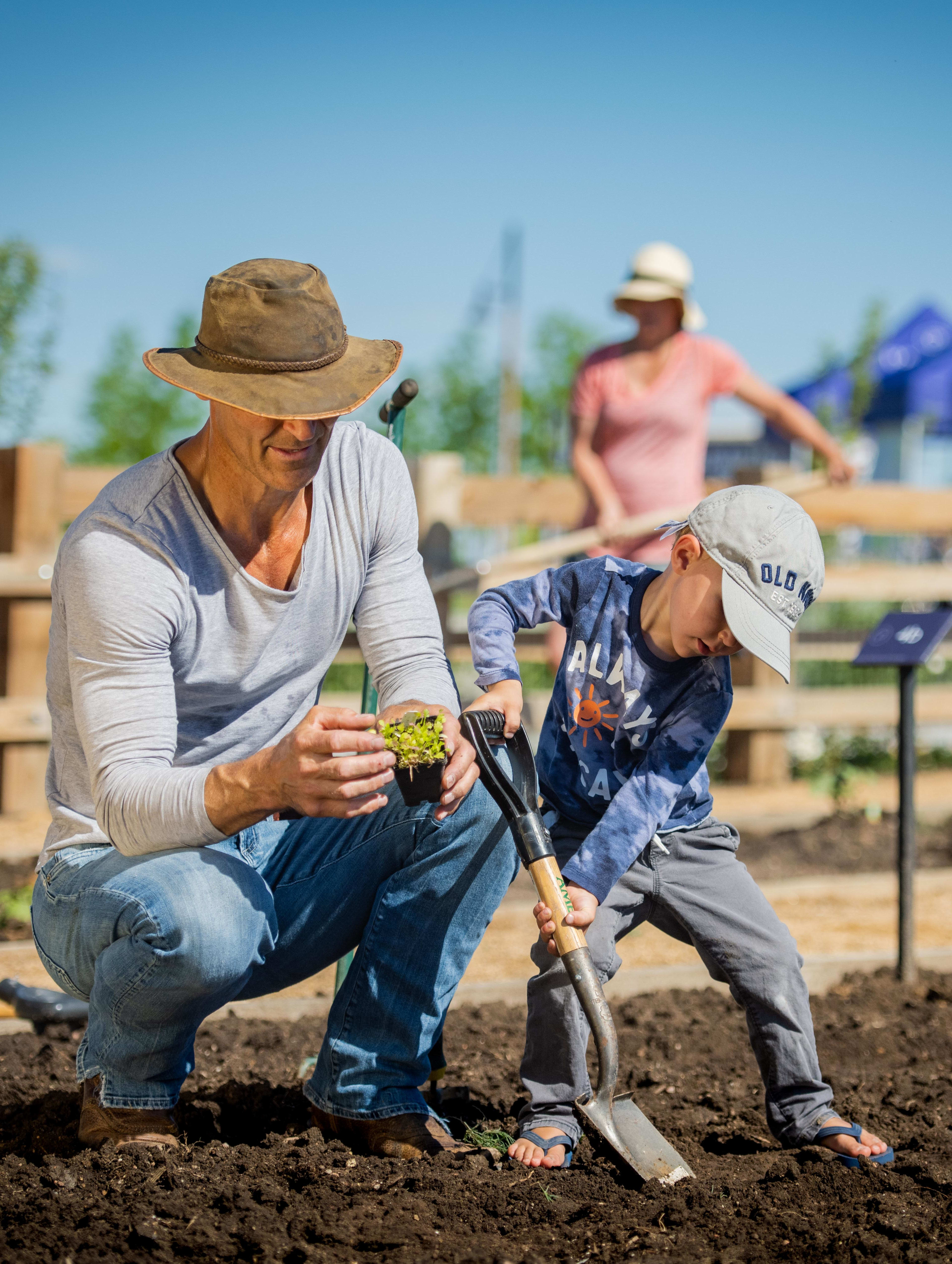 A person in casual clothing is working in a garden plot, with a person in the background wearing a pink shirt.