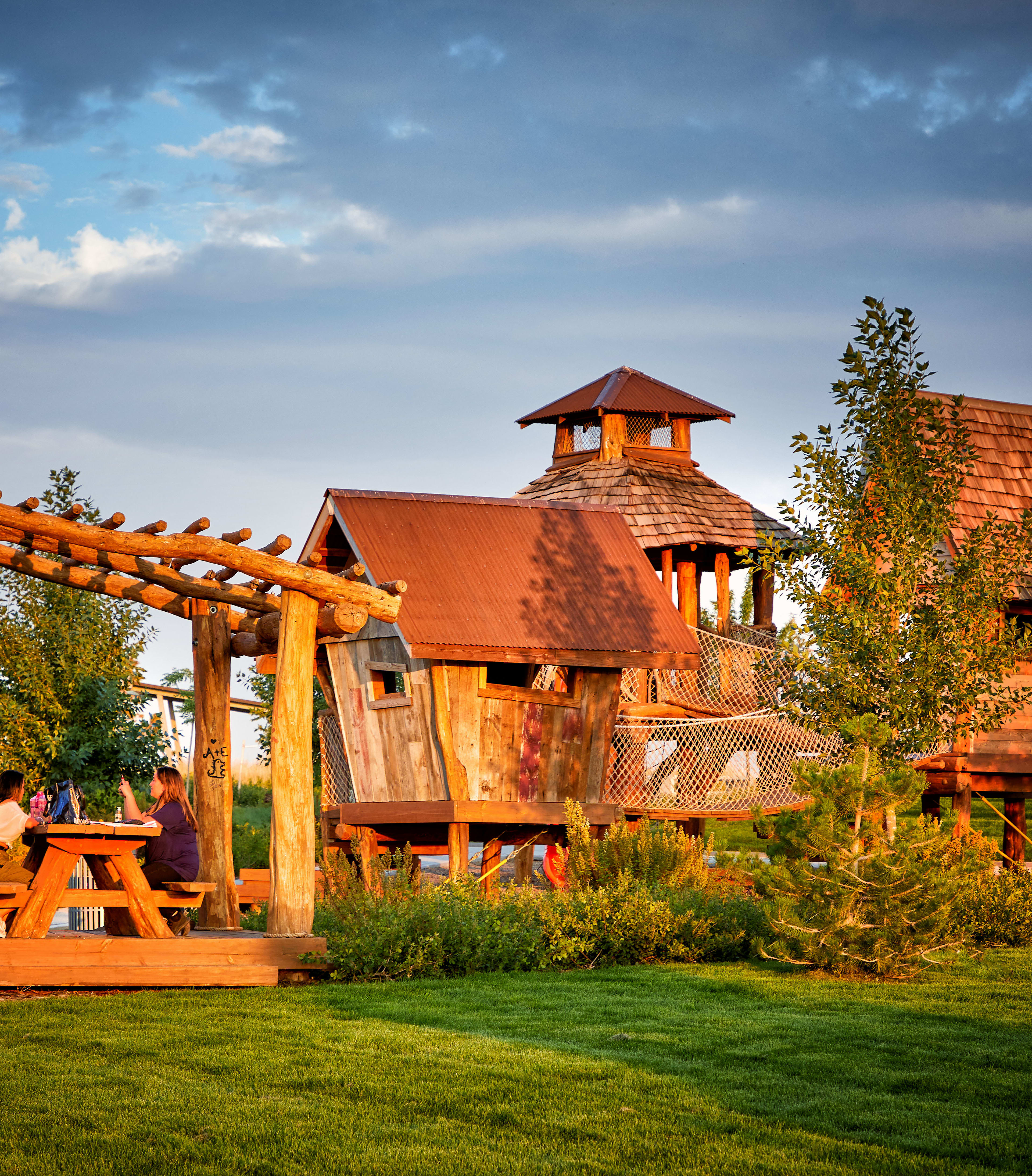 A rustic wooden structure with a thatched roof stands in a lush green garden, surrounded by trees and a cloudy sky in the background.