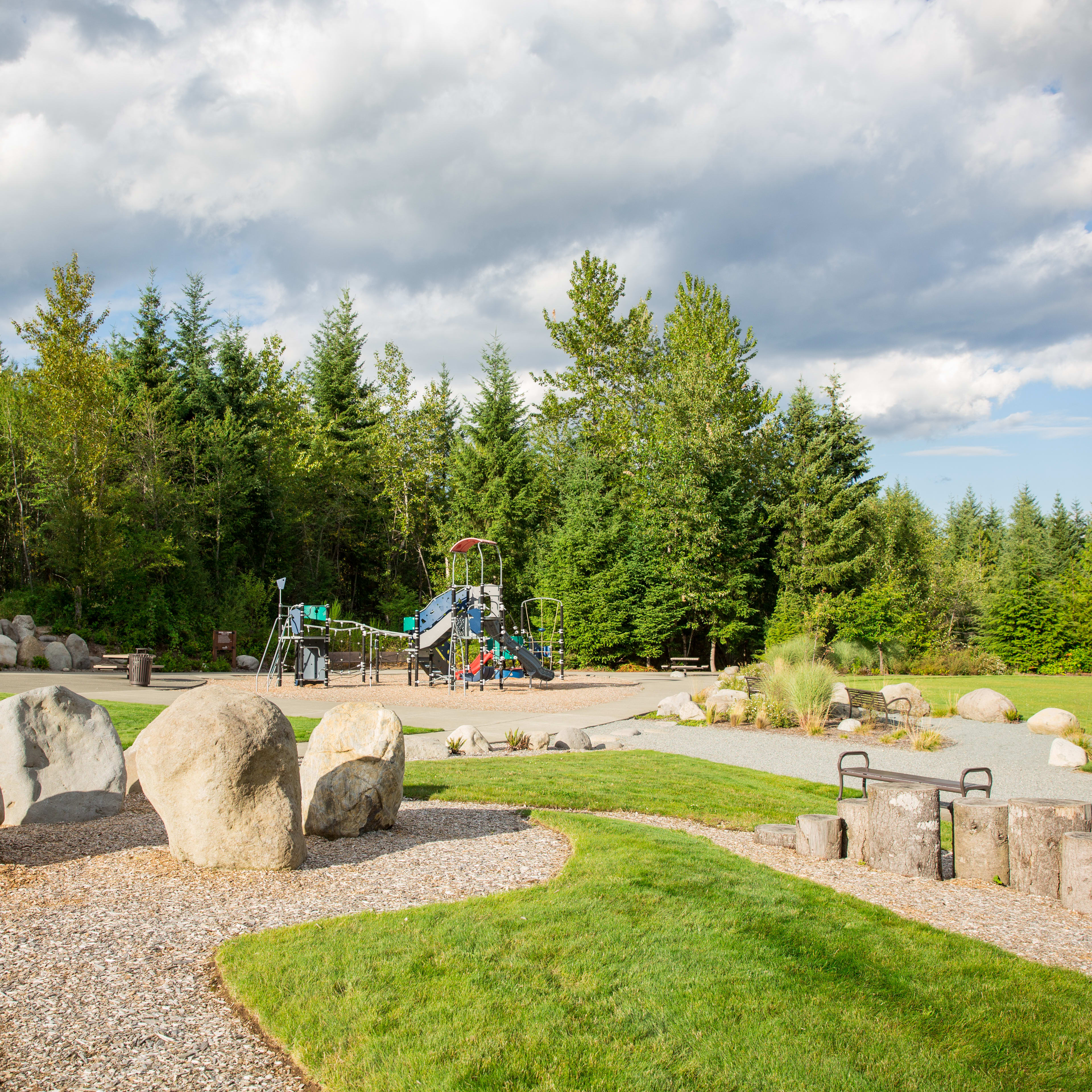 A lush, green park with a playground and stone structures surrounded by a dense forest of pine trees under a cloudy sky.