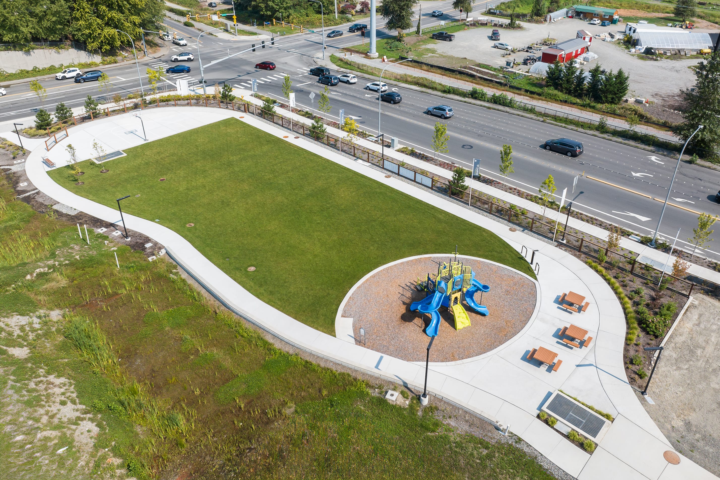 A grassy park with a colorful playground structure surrounded by a paved walkway, with a busy road and traffic visible in the background.