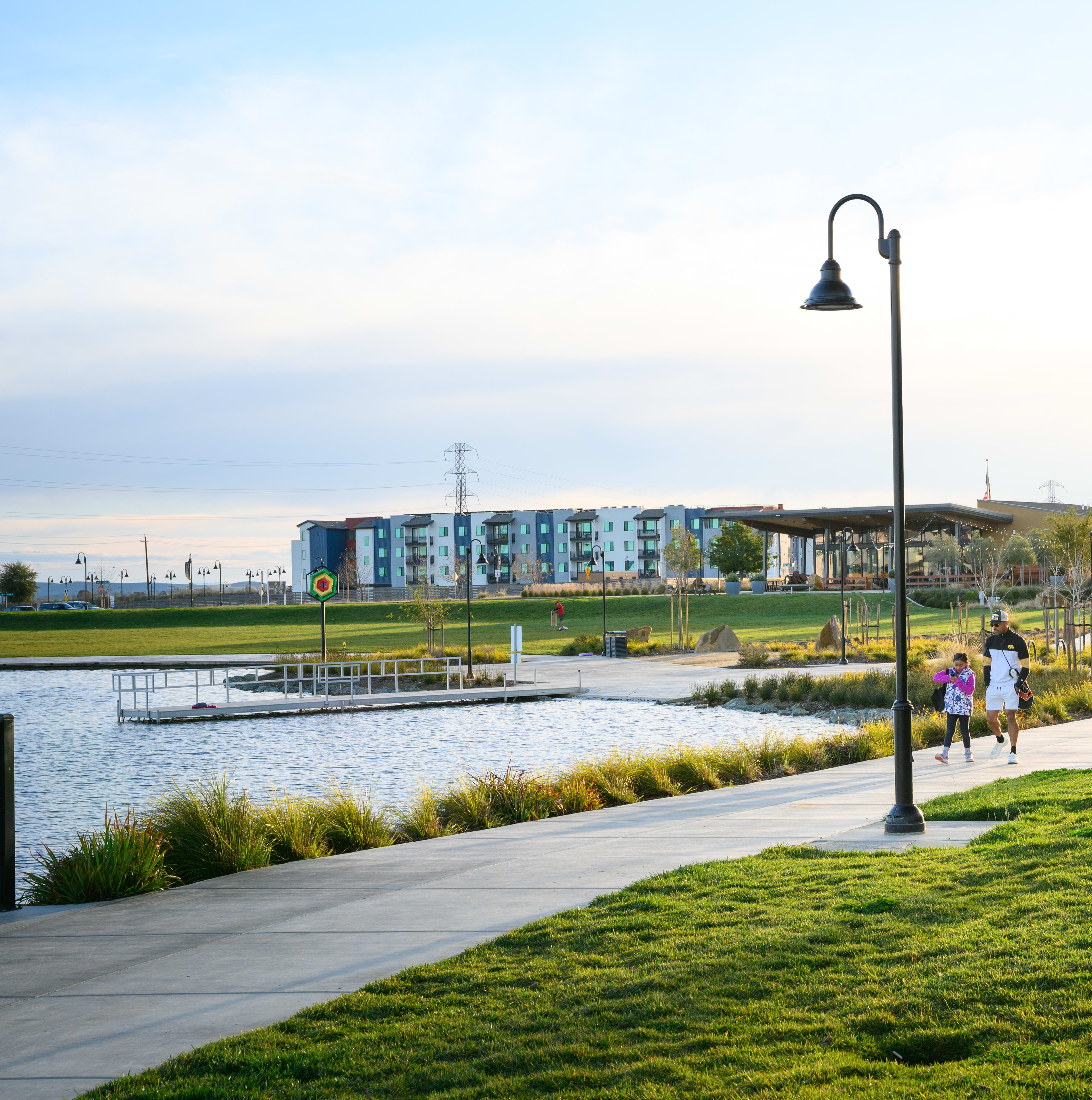 A scenic outdoor public space with a paved walkway, lush green grass, and a decorative lamp post, with a body of water and colorful buildings visible in the background.
