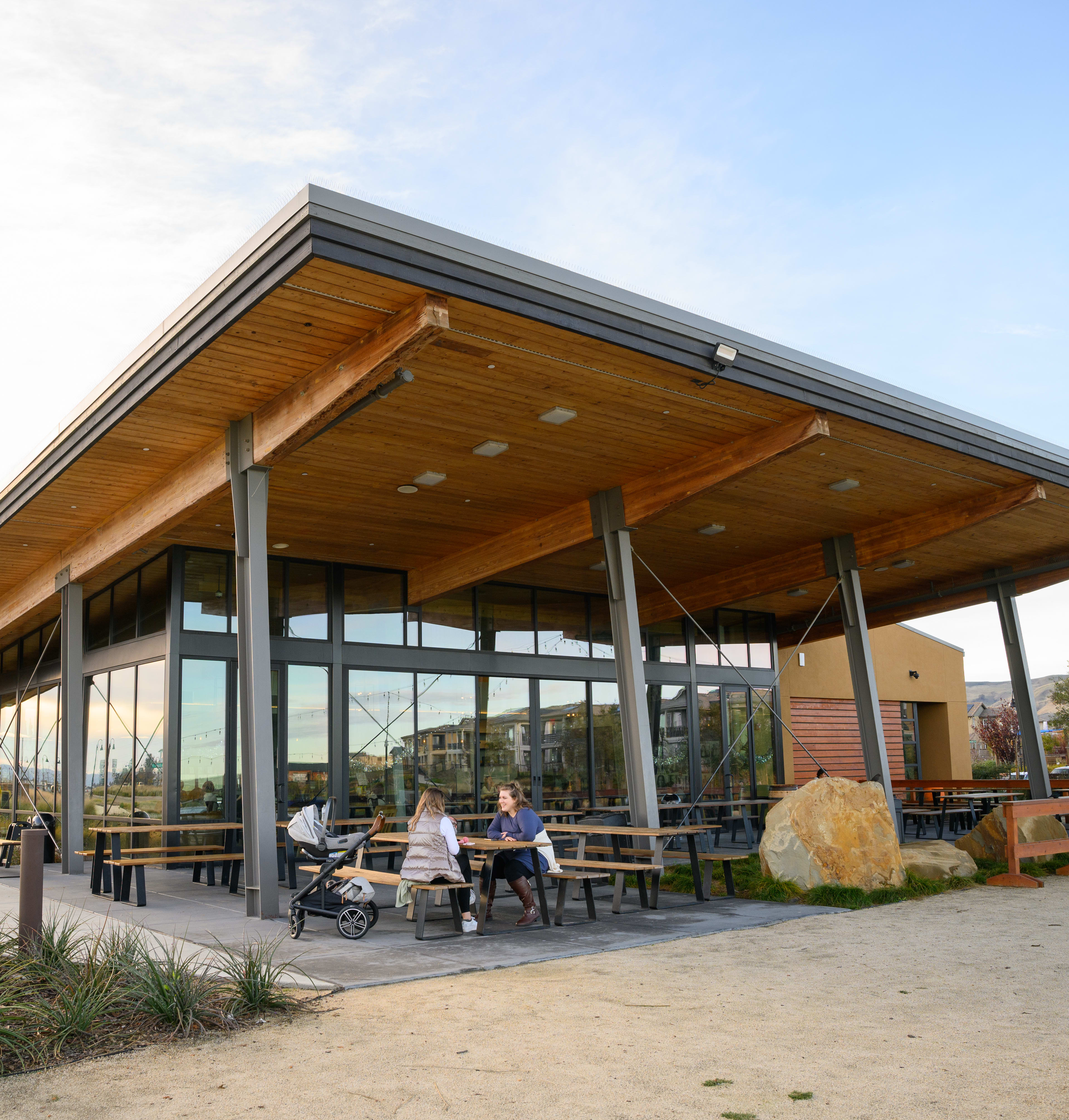 A modern, wooden-framed building with a large covered patio area, surrounded by landscaping and outdoor seating, set against a backdrop of a clear sky.