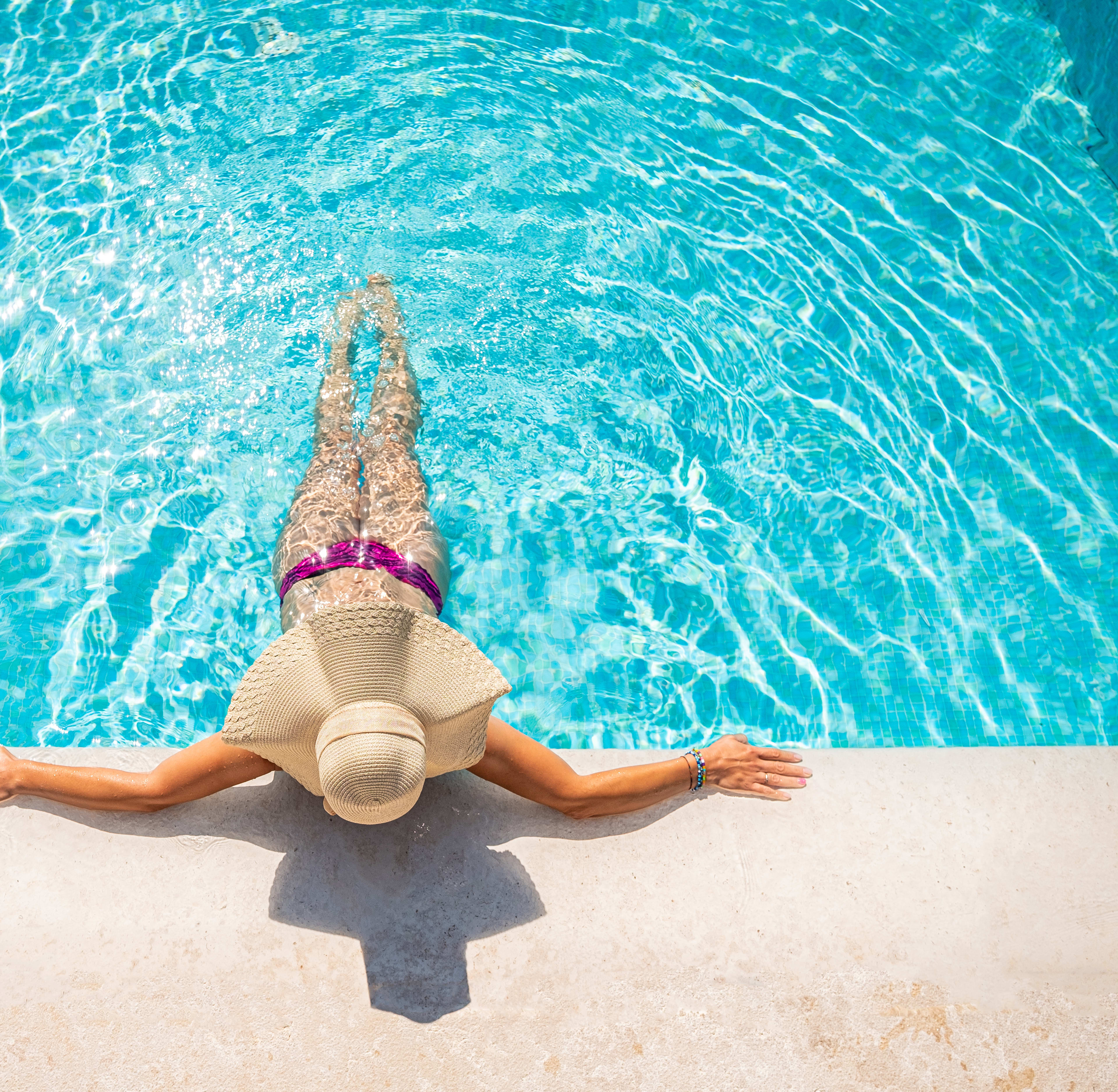 A person in a pink swimsuit is relaxing on the edge of a clear, turquoise swimming pool, with their legs dangling in the water.
