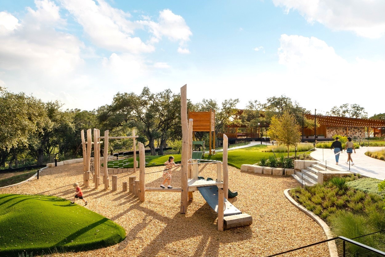 A well-designed outdoor playground with wooden structures, grassy areas, and walking paths surrounded by lush greenery and a clear sky.