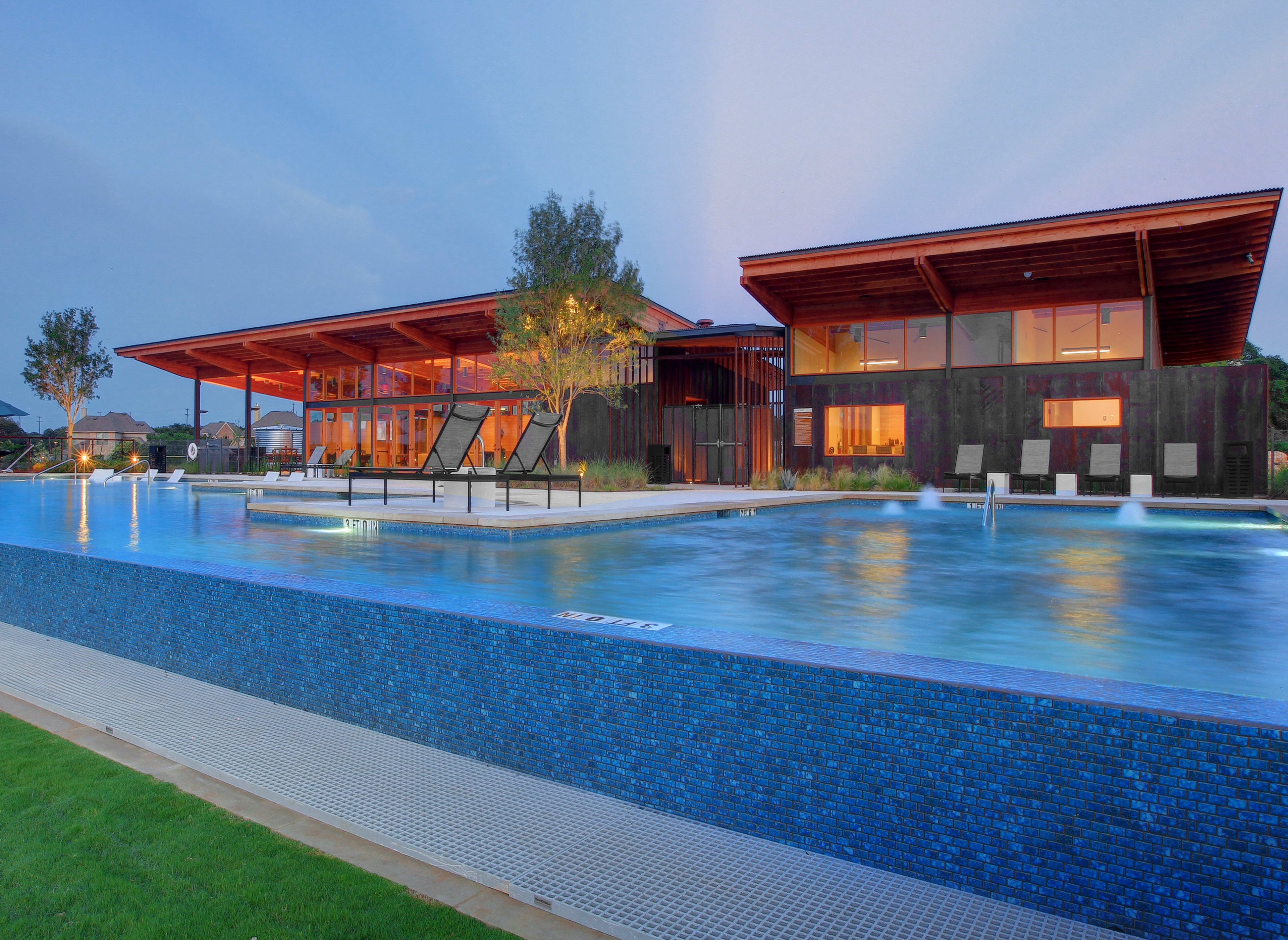 A modern, multi-story building with a large swimming pool in the foreground, surrounded by lush greenery and a clear blue sky.