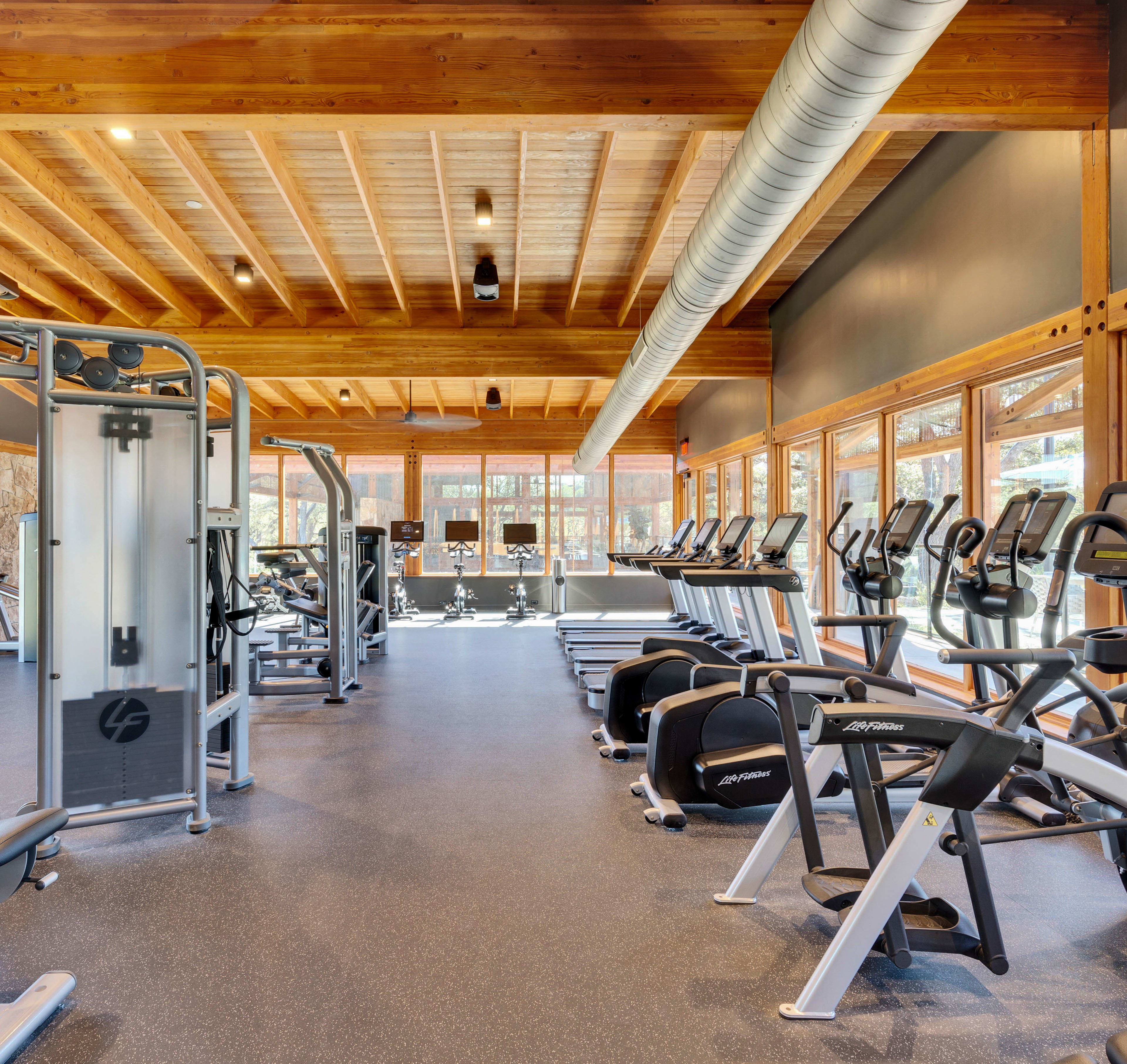 A well-equipped gym with various exercise equipment, including treadmills, elliptical machines, and weight training machines, set against a backdrop of wooden beams and large windows providing natural light.