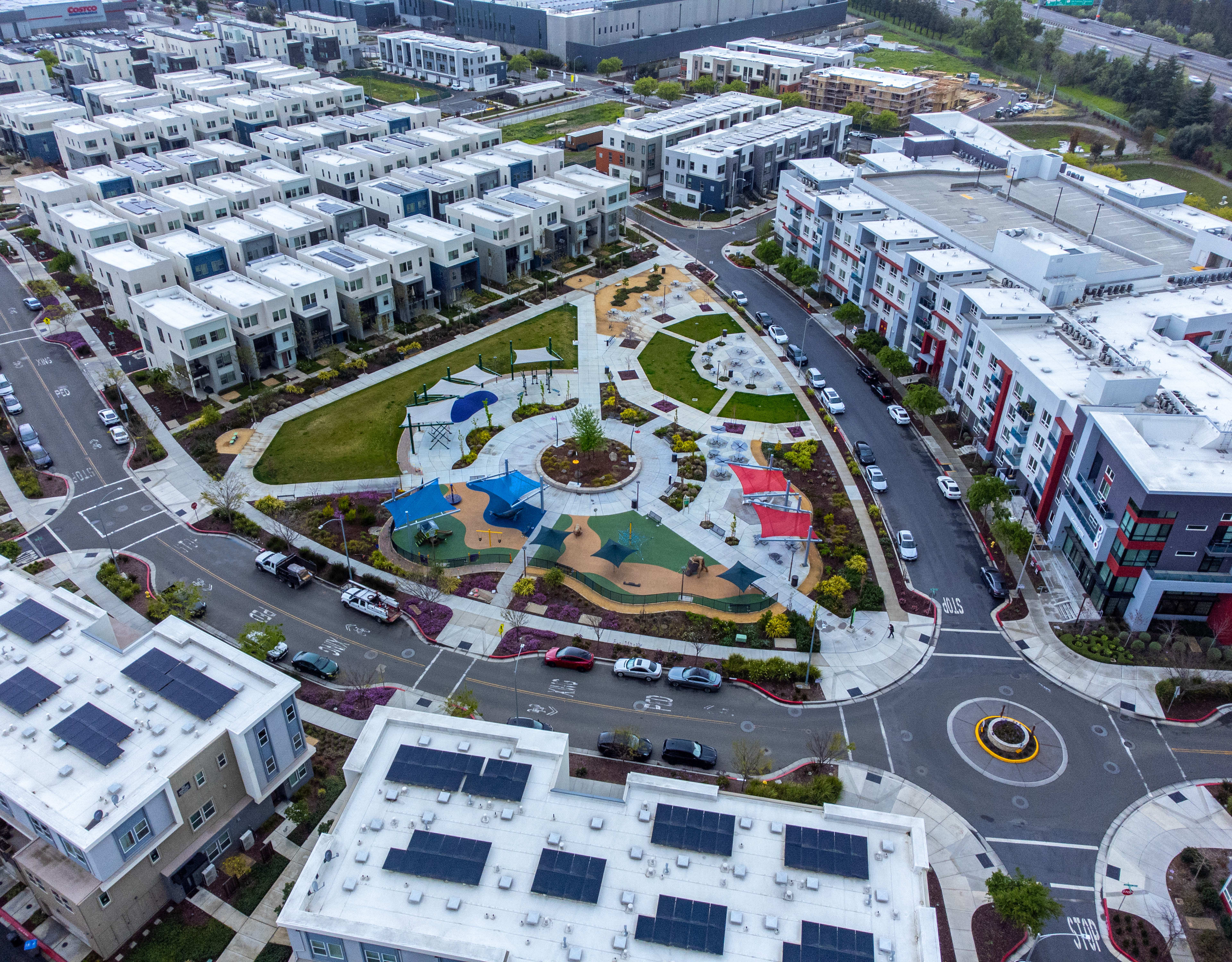 A vibrant and well-designed urban landscape with a central park-like area featuring colorful landscaping, walkways, and buildings surrounding it.