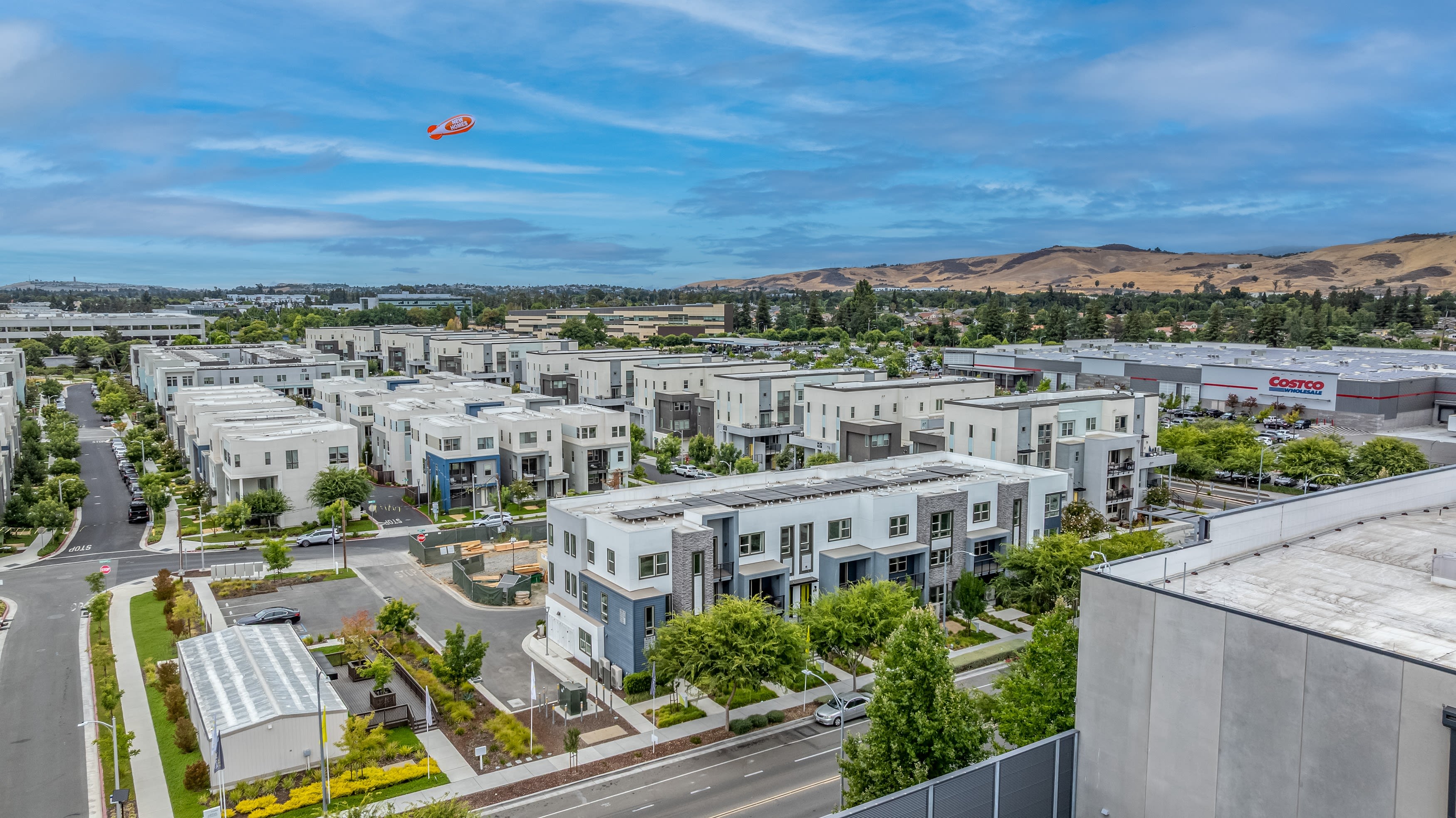 A densely populated urban neighborhood with rows of white apartment buildings surrounded by lush greenery, set against a backdrop of rolling hills and a bright blue sky with a hot air balloon floating overhead.