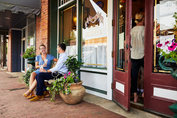 Two people are sitting on a bench outside a brick building, surrounded by potted plants and a wooden door.