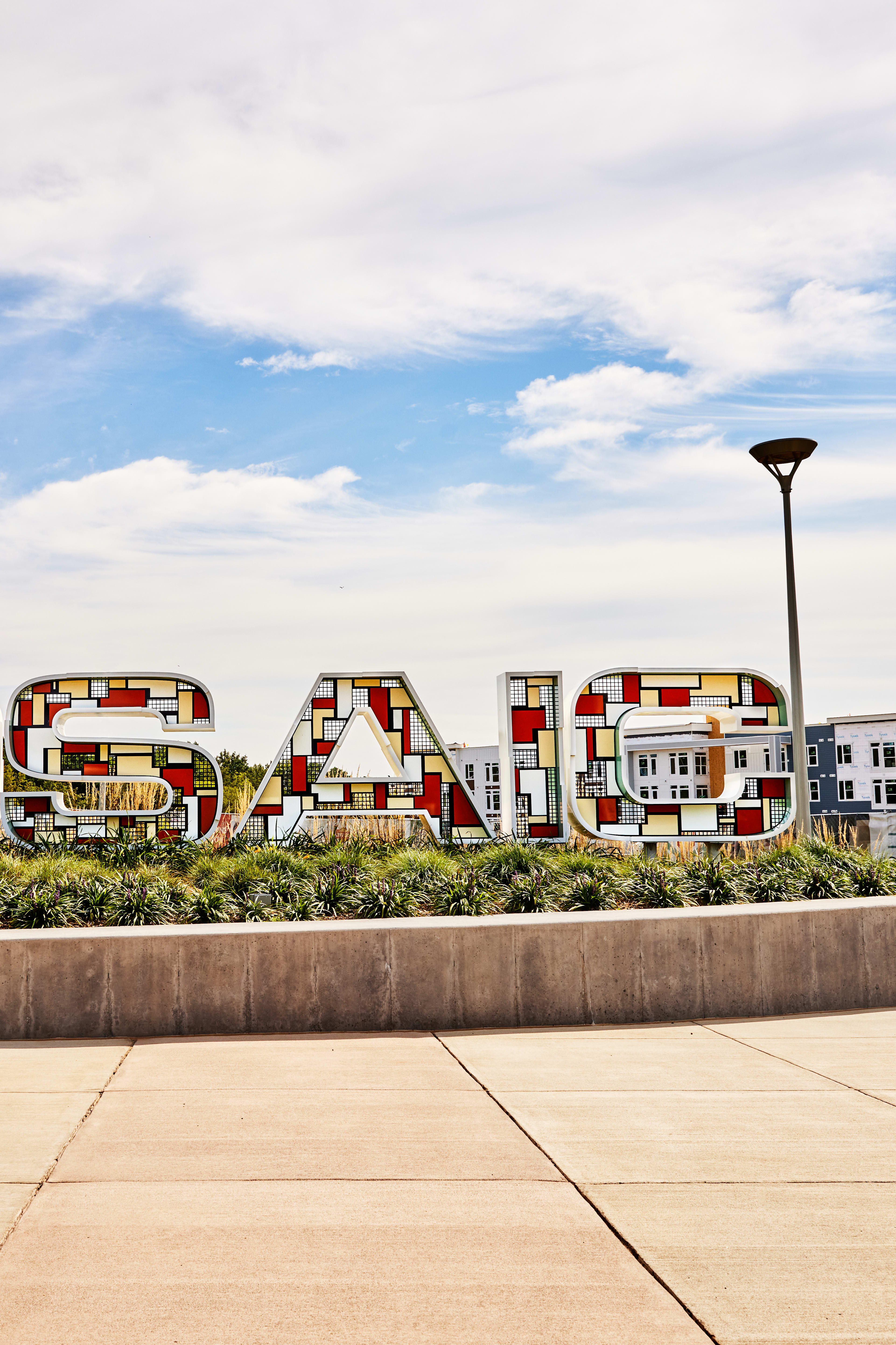 A large, colorful "Mosaic" sign stands in the foreground, surrounded by a paved plaza and landscaping, with a cloudy blue sky and buildings visible in the background.