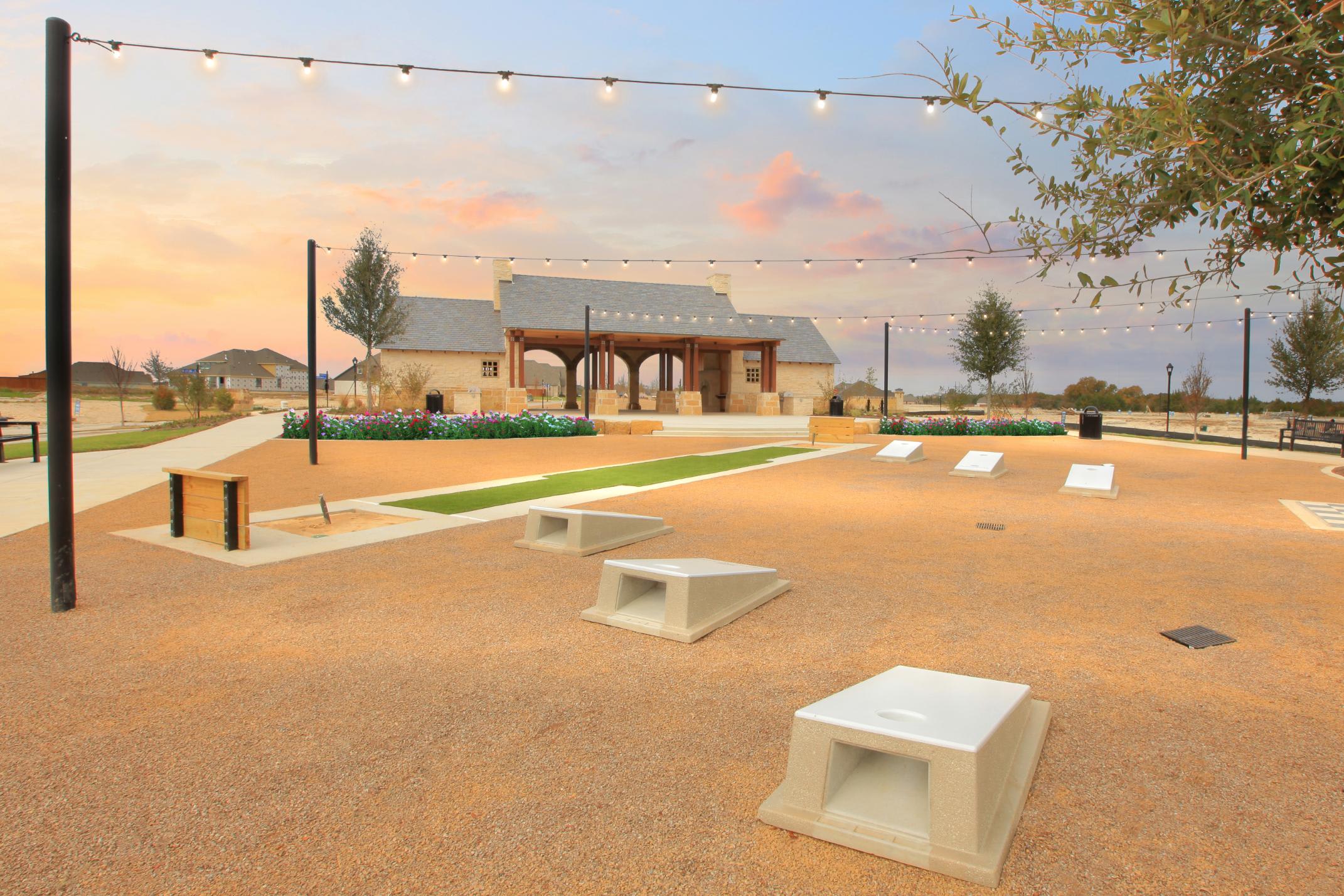 A public outdoor space with a wooden structure, string lights, and benches set against a backdrop of trees and a colorful sky.