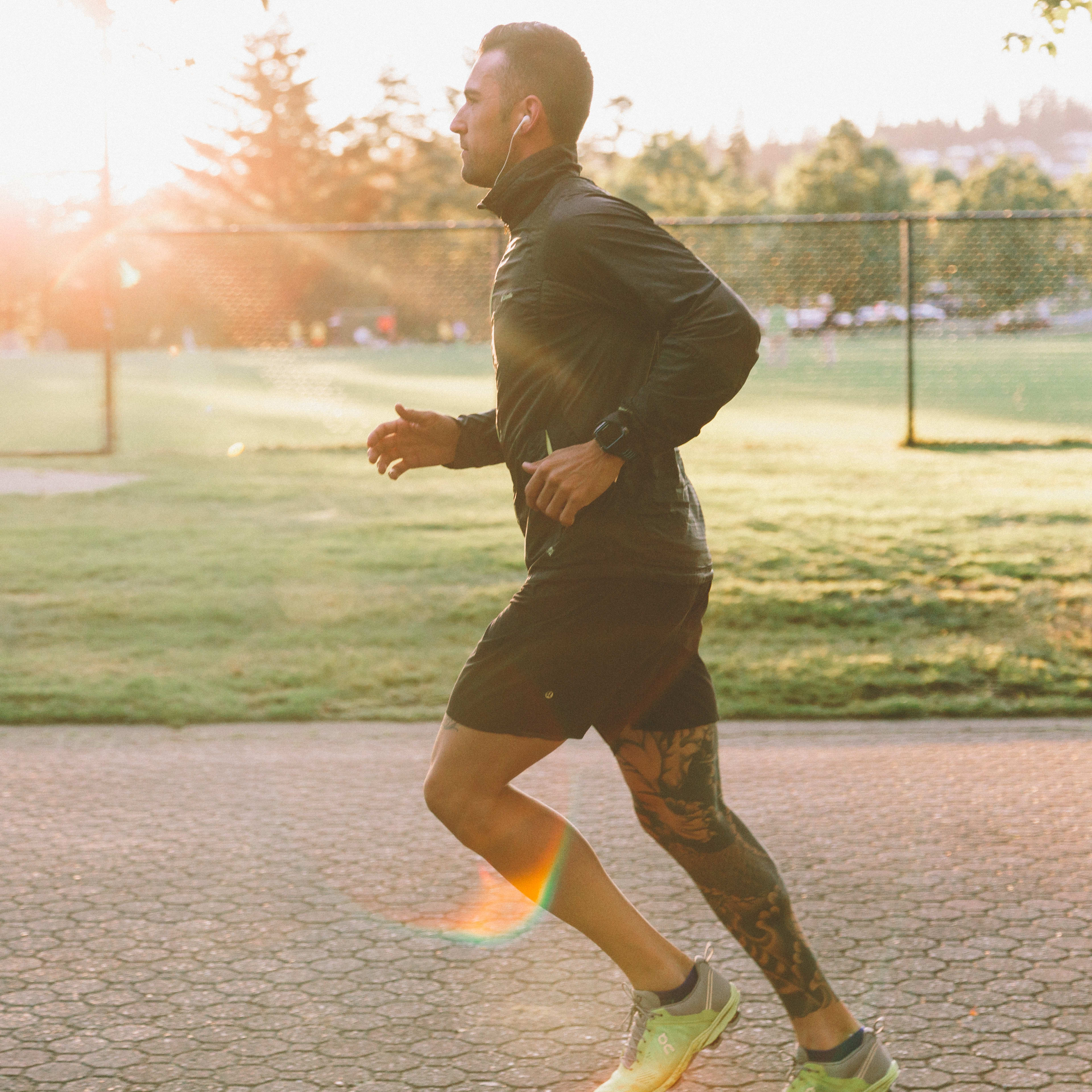 A man in athletic clothing is jogging on a paved path surrounded by a grassy field and trees, with the sun shining brightly in the background.