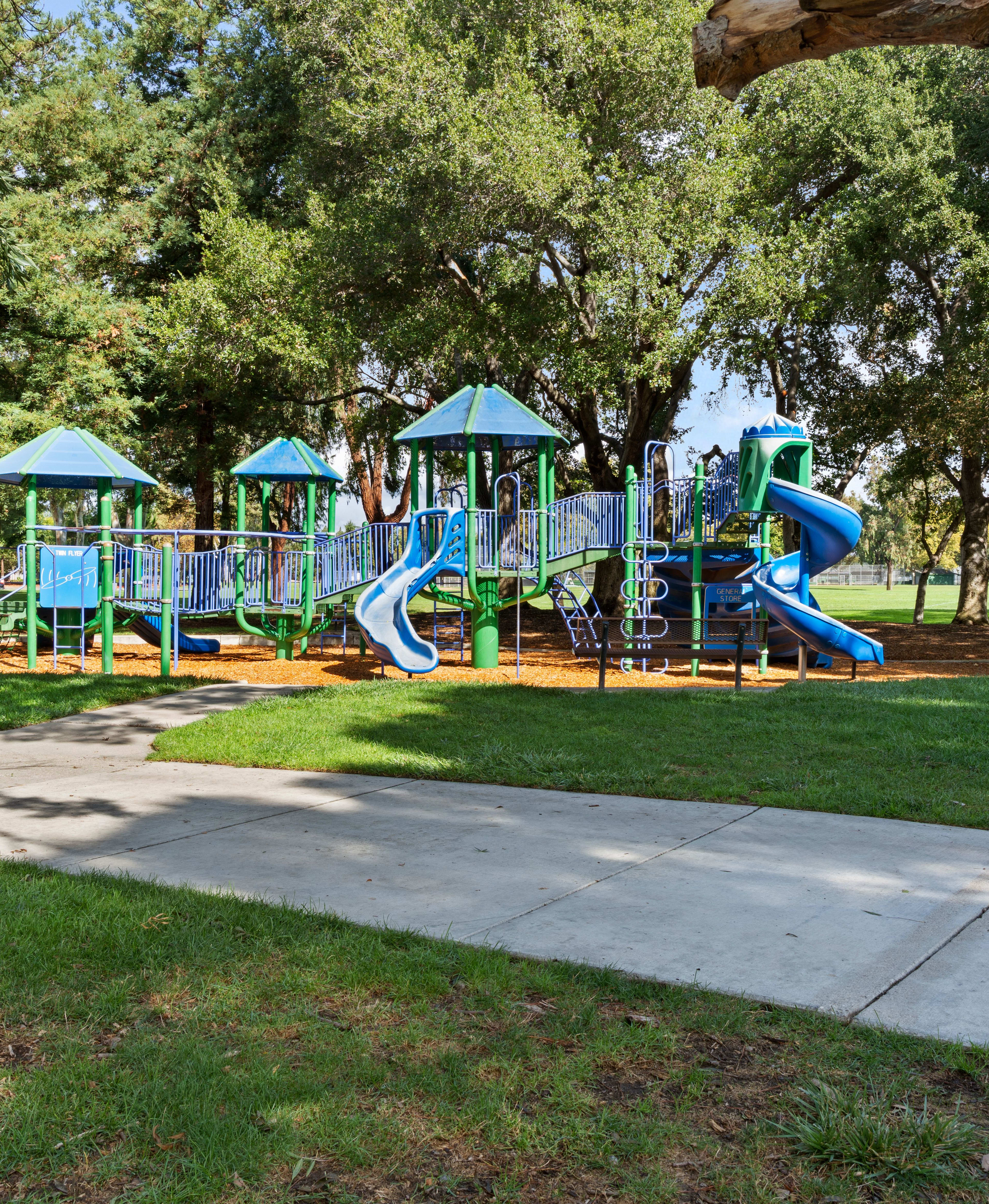 A colorful playground with various play structures, surrounded by lush greenery and trees, sits in a well-maintained park setting.