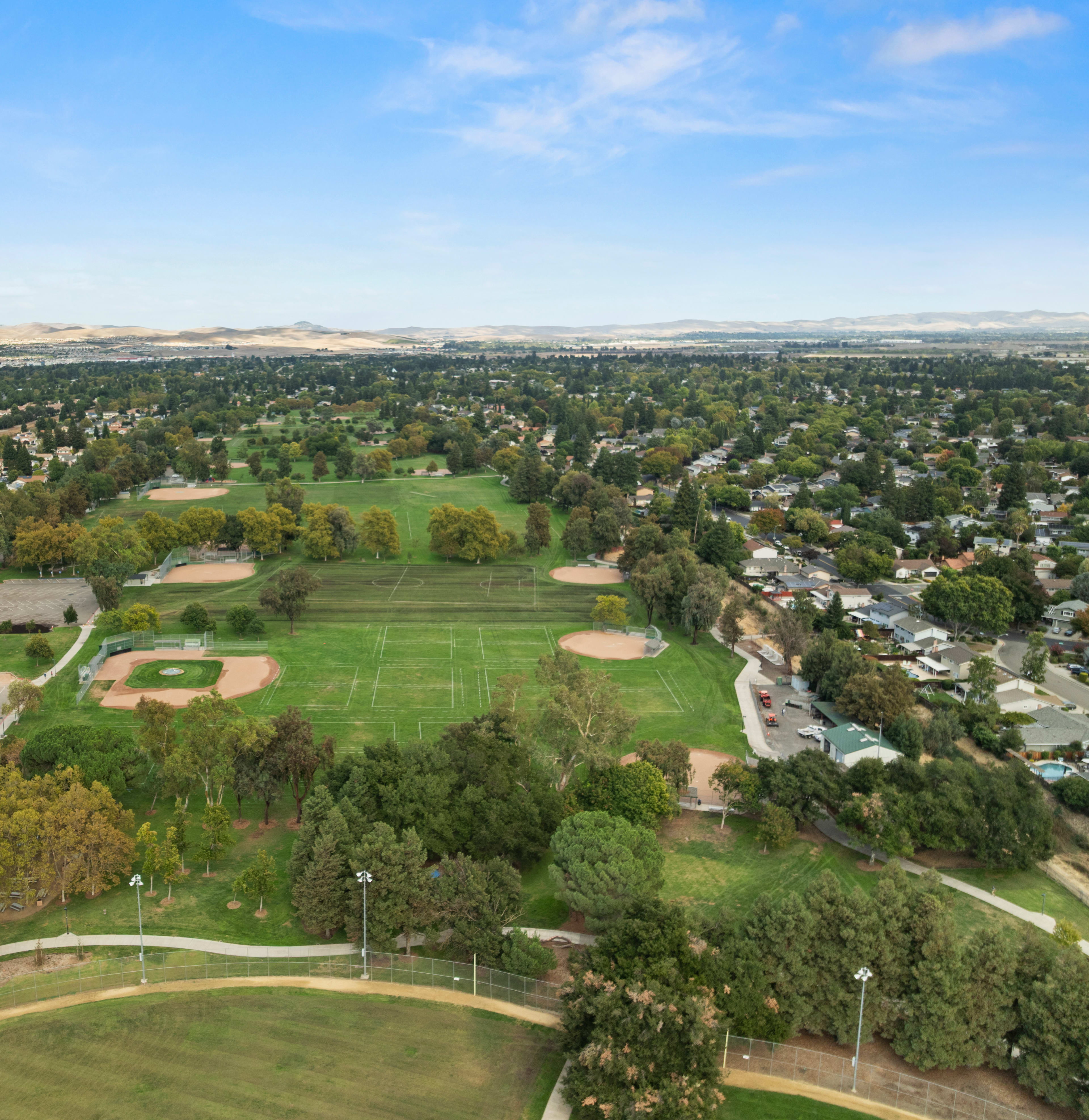 An aerial view of a lush, green park surrounded by a residential area, with mountains visible in the distance under a clear blue sky.