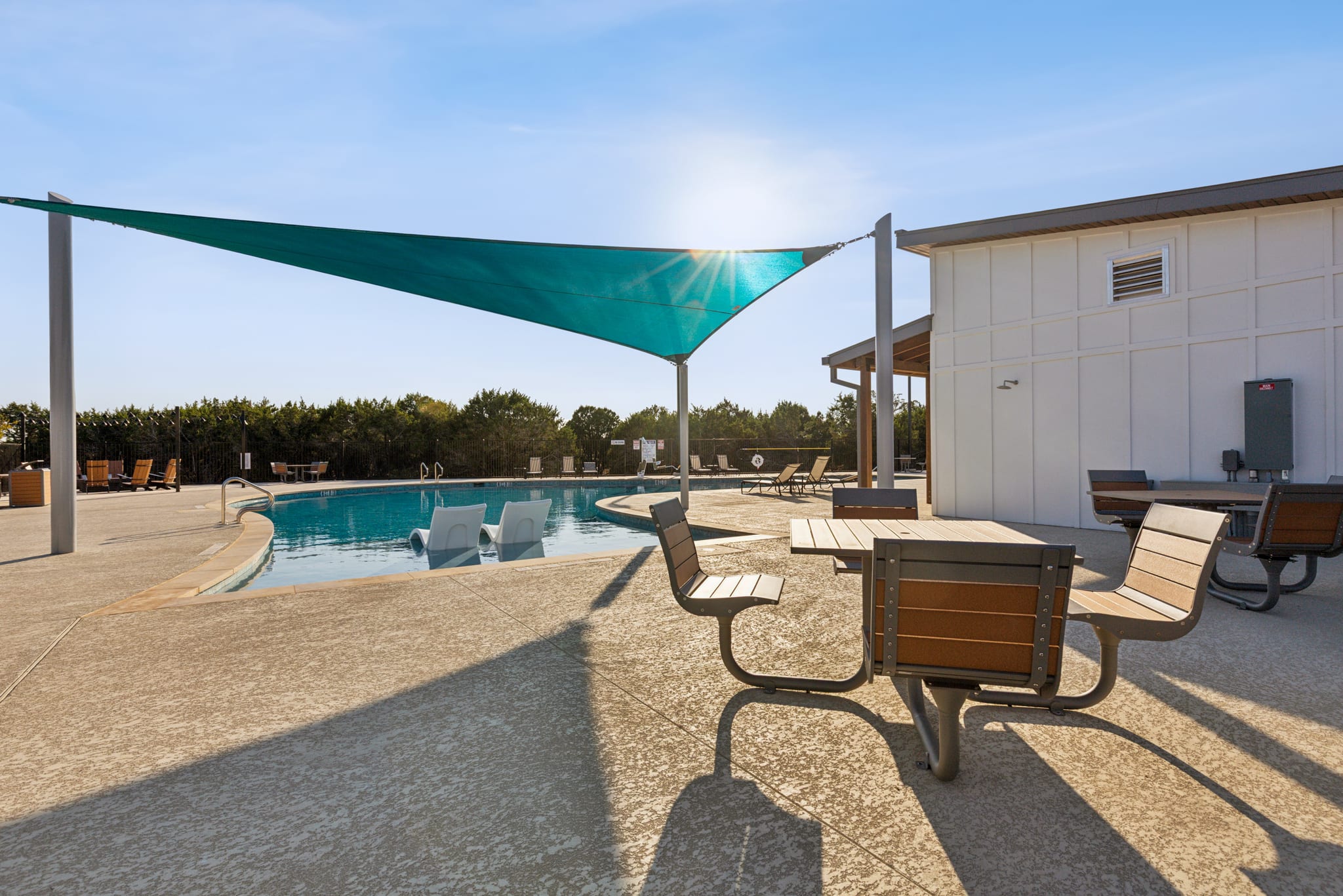 A swimming pool surrounded by lounge chairs and a large green shade sail, with a building in the background and a bright, sunny sky overhead.