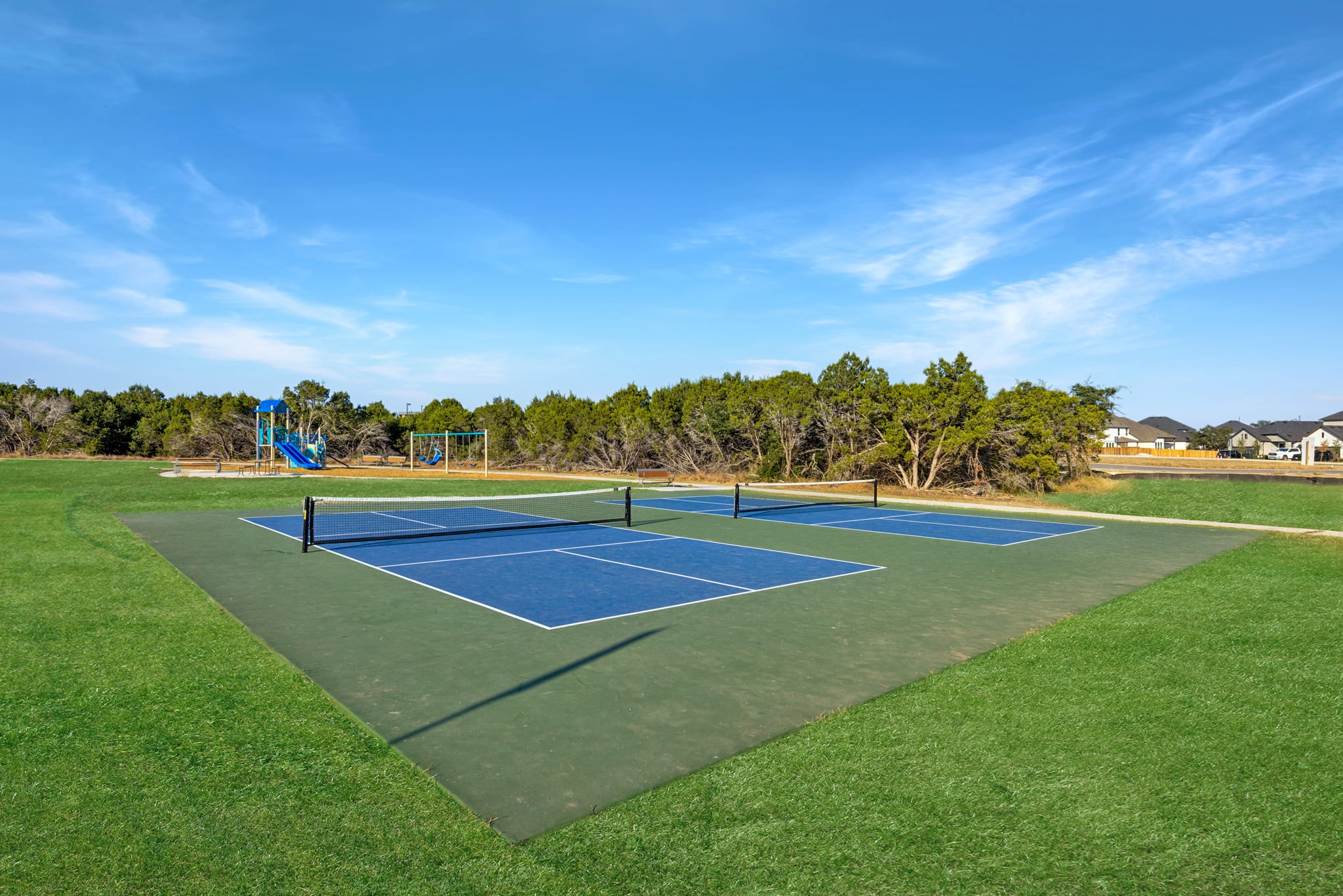 A well-maintained tennis court with a blue playing surface surrounded by a lush green lawn, set against a backdrop of a blue sky with wispy clouds and a forested area.