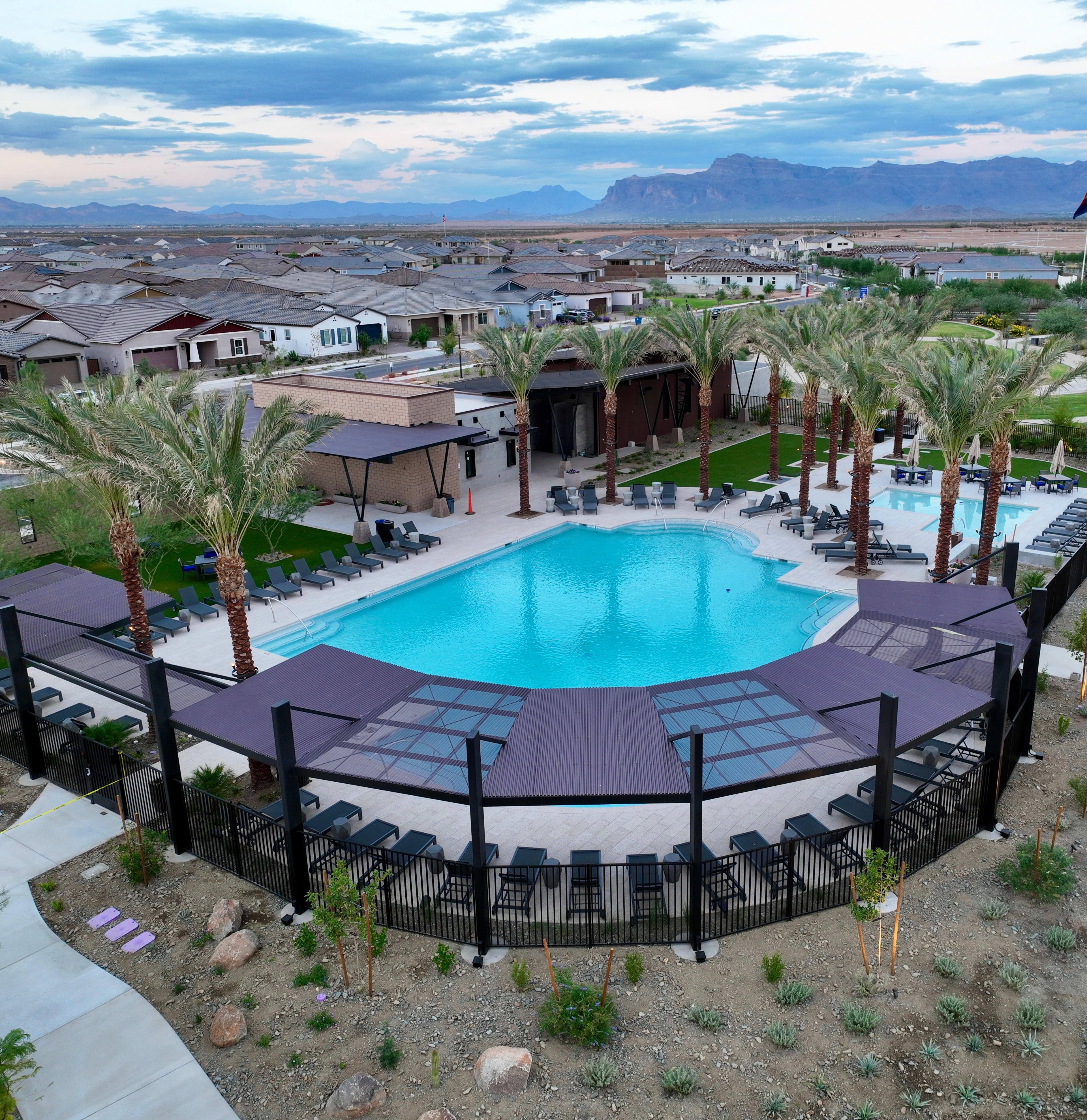 A large outdoor swimming pool surrounded by palm trees and buildings, with a mountainous landscape visible in the background.