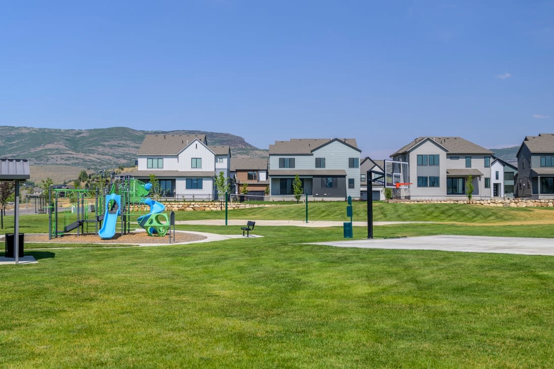 A well-manicured grassy park with colorful playground equipment in the foreground, surrounded by a residential neighborhood with modern, multi-story homes in the background.