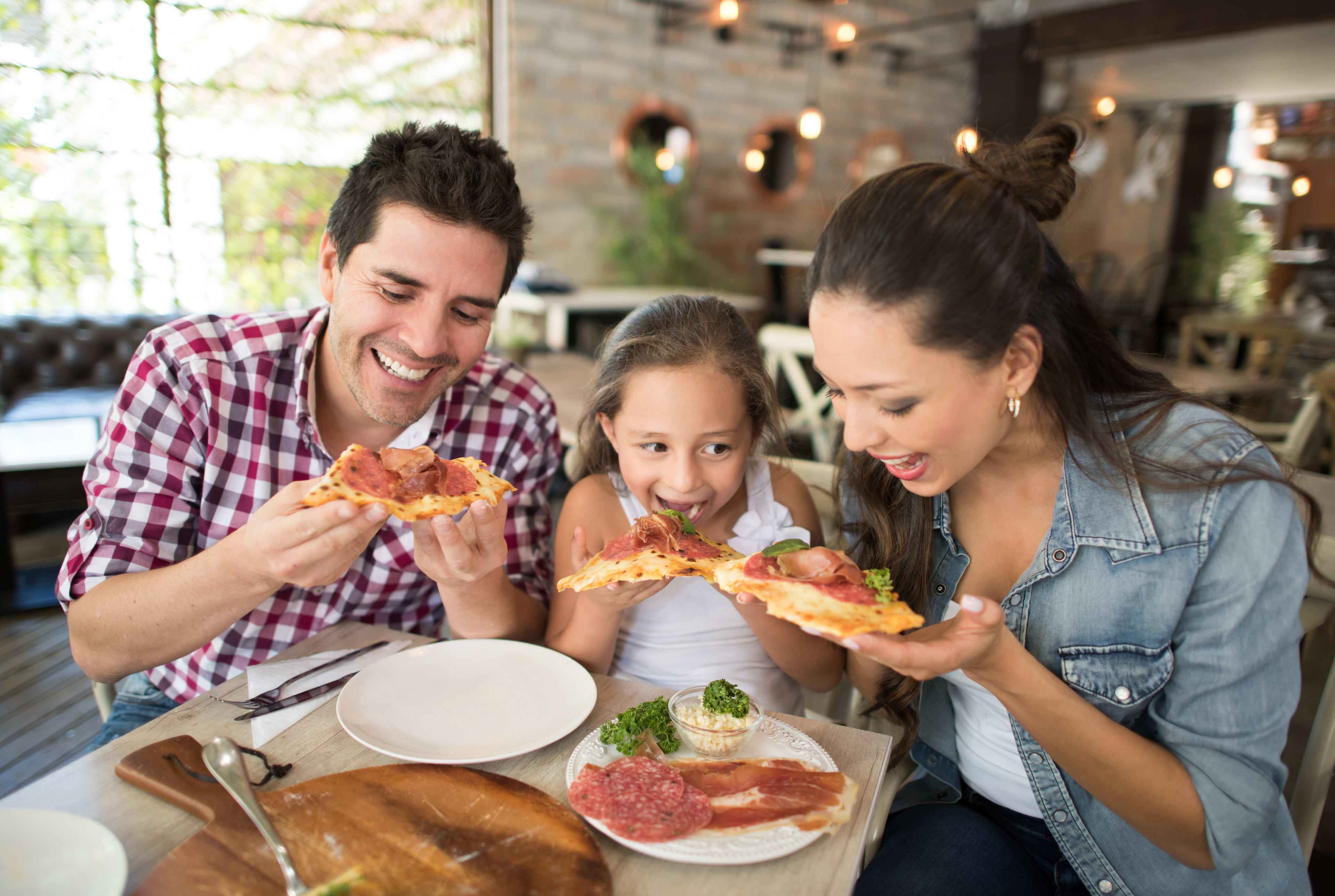 A family of three enjoying a meal together, with the adults and child all eating slices of pizza in a cozy, well-lit restaurant setting.