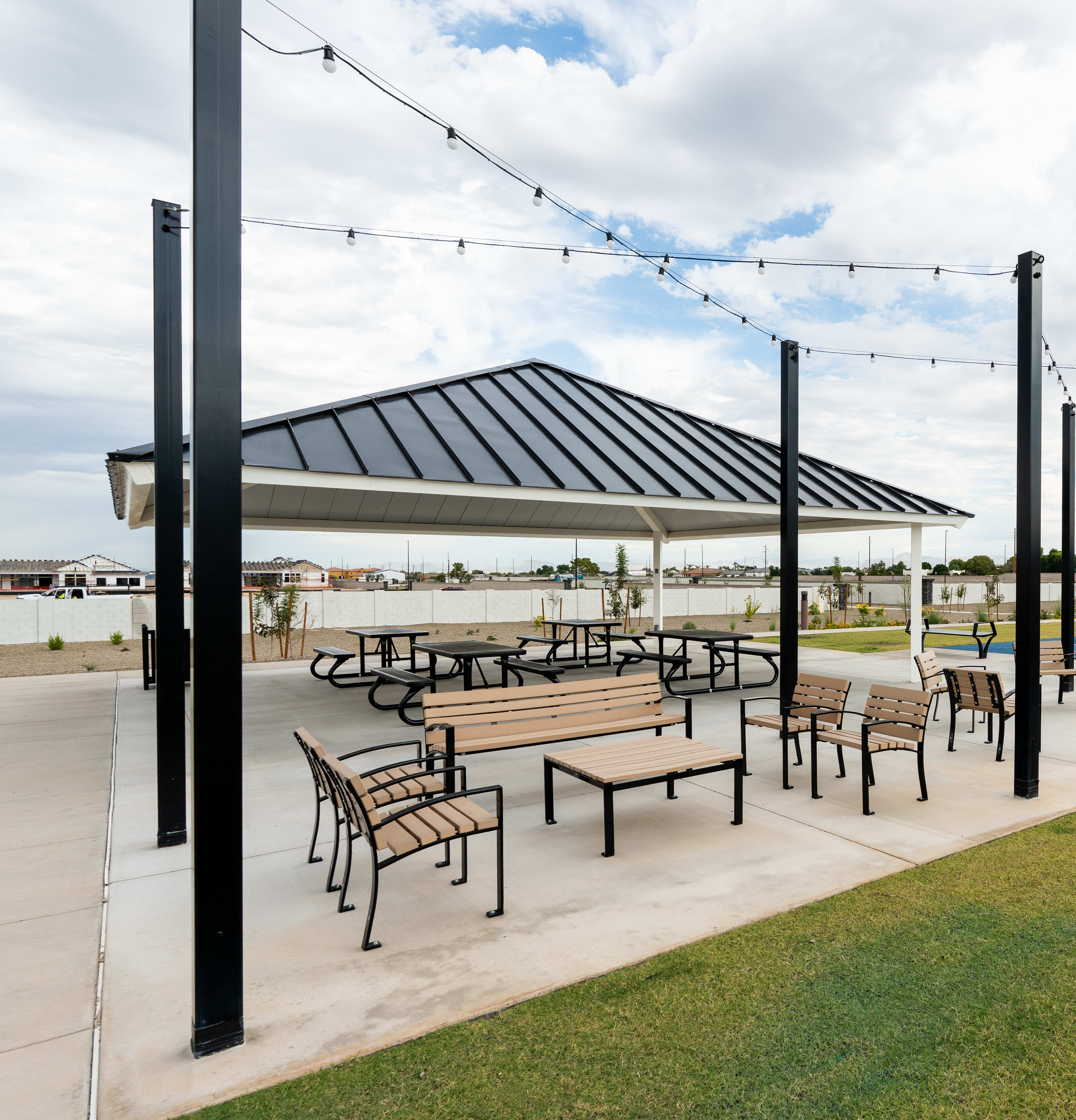 A covered outdoor seating area with tables and chairs, surrounded by a grassy lawn and a swimming pool in the background, with string lights overhead and a cloudy sky above.