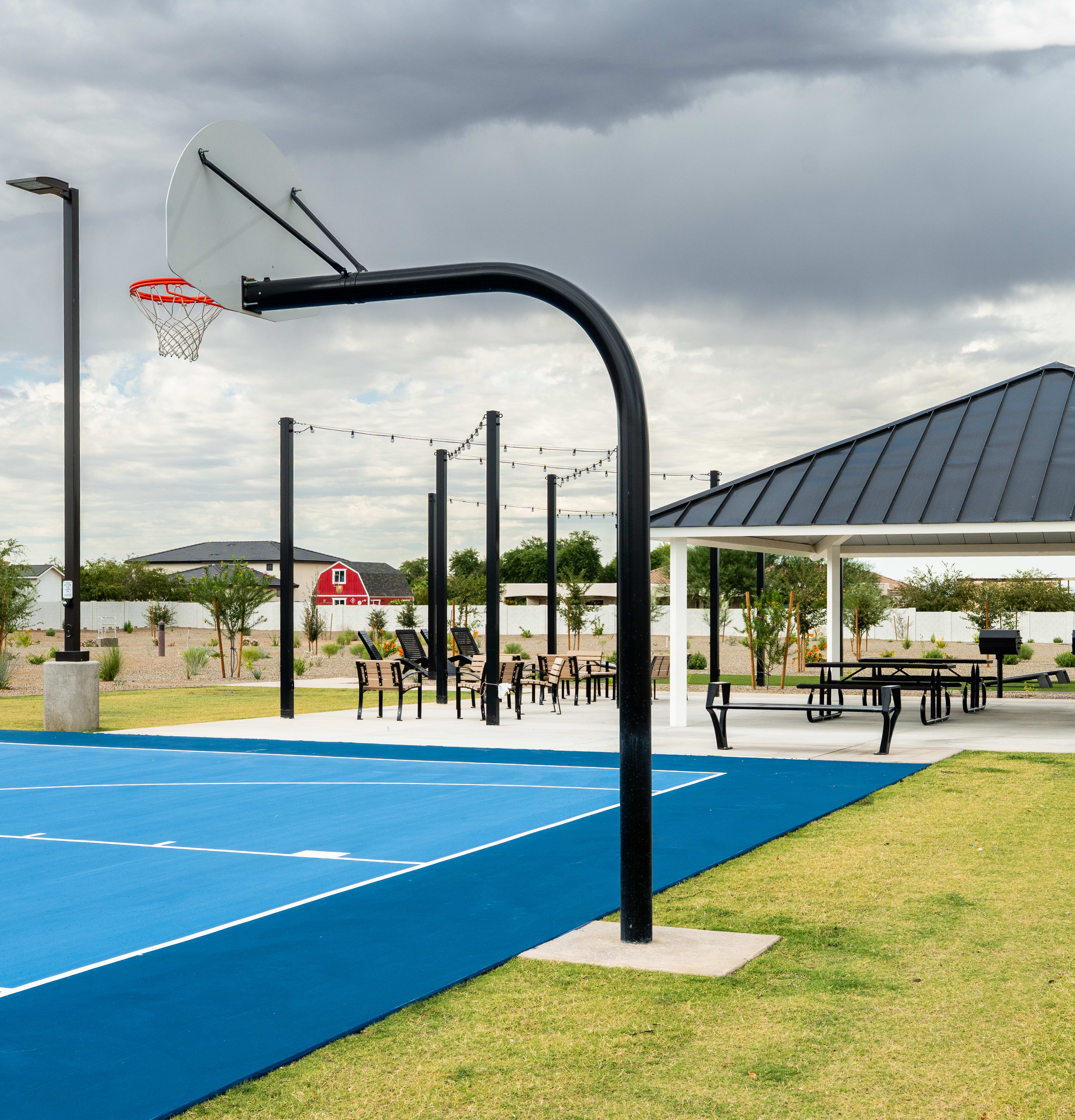 A basketball court with a hoop and net stands in the foreground, surrounded by a grassy area and a covered pavilion structure in the background against a cloudy sky.