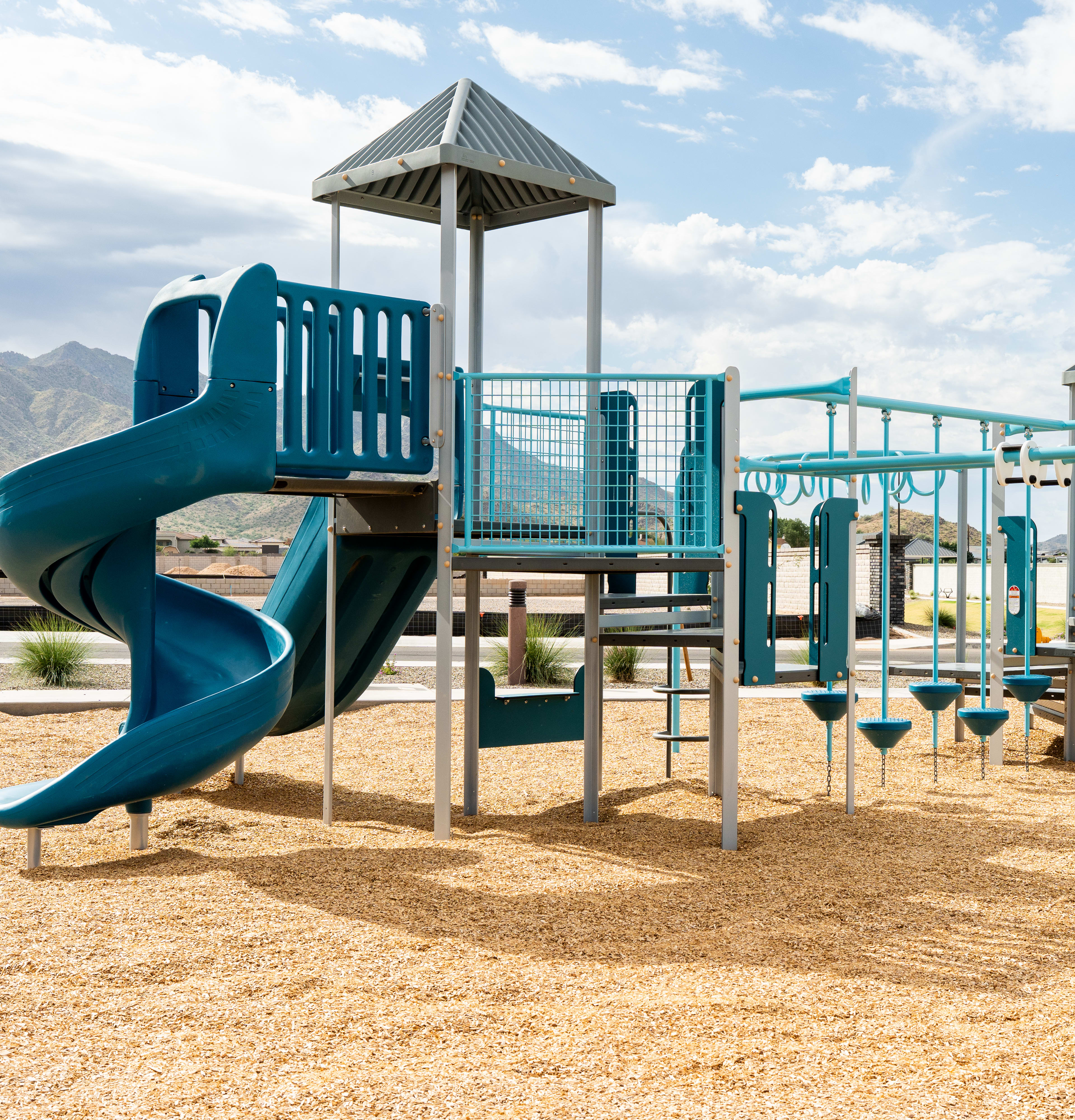 A colorful and modern playground structure with slides, ladders, and platforms stands in a sandy area, surrounded by a mountainous landscape under a cloudy sky.