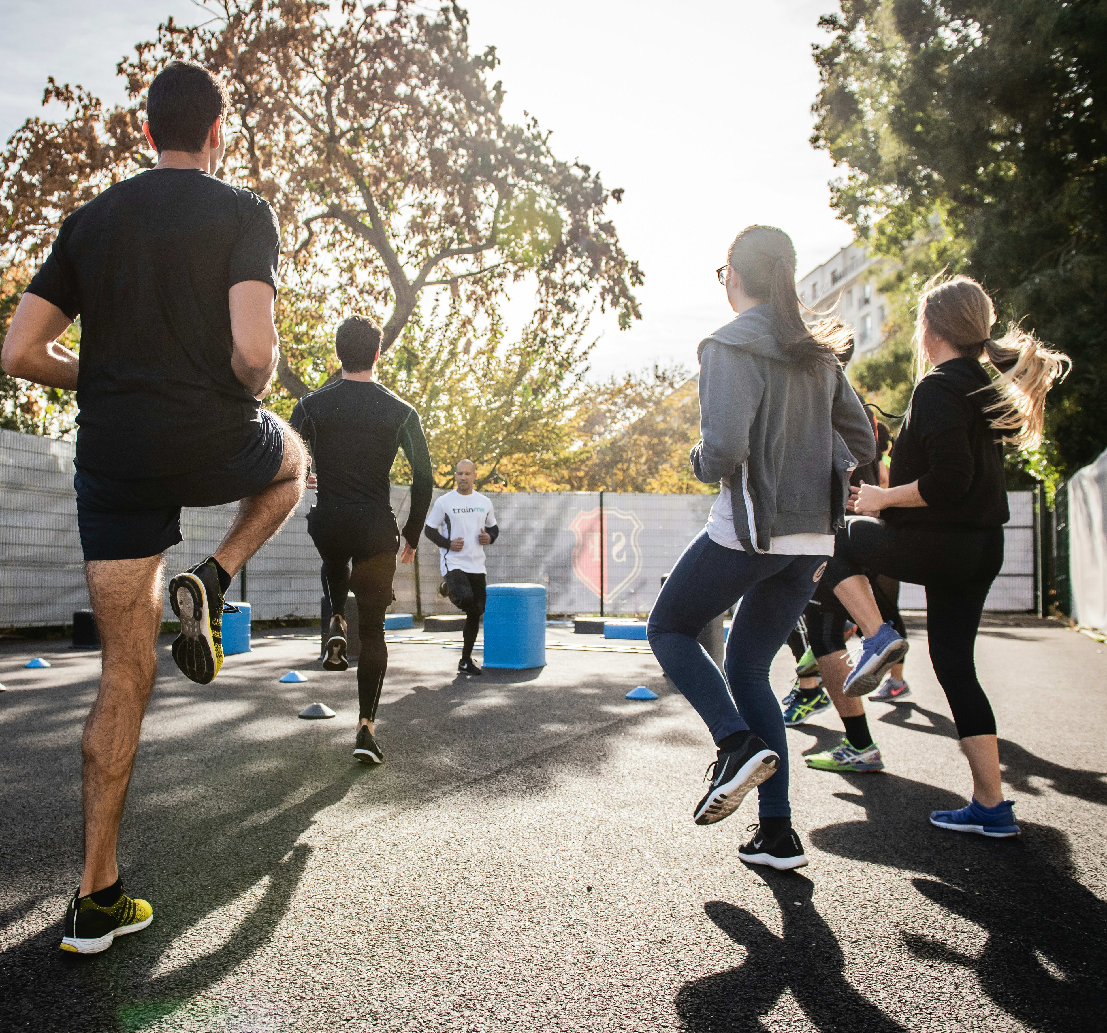 A group of people, some wearing athletic clothing, are walking or running on a paved path surrounded by trees and buildings in the background.