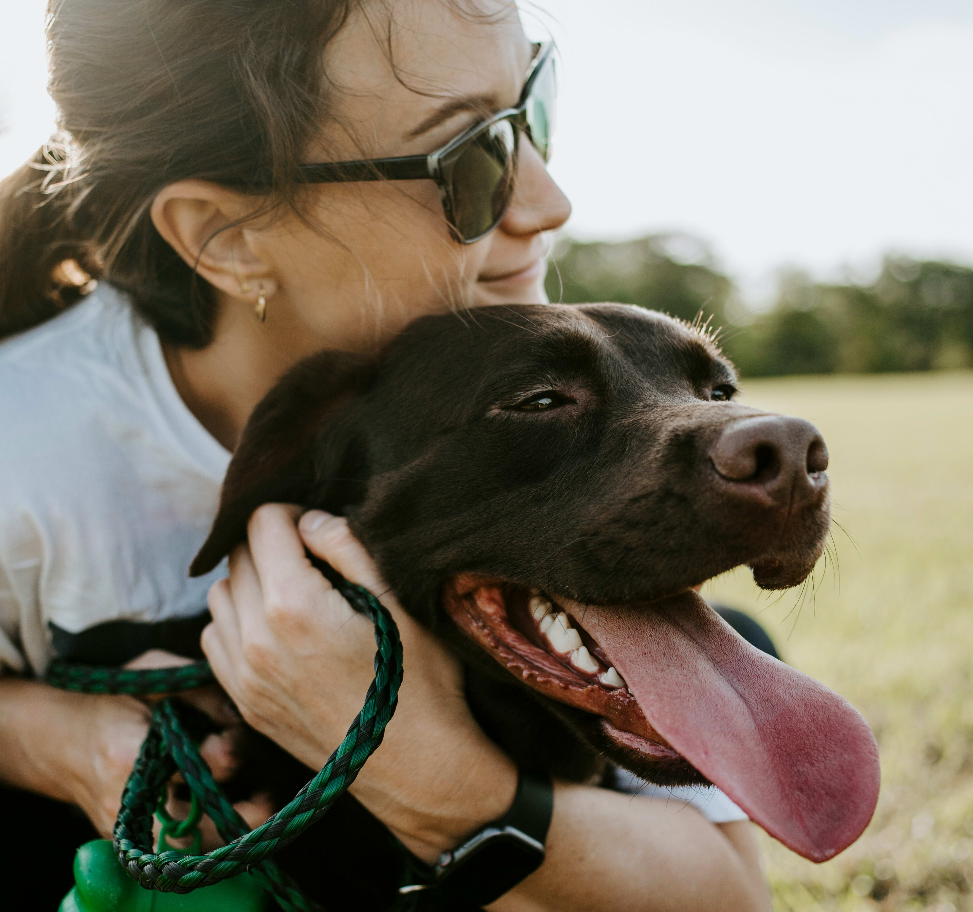 A woman in sunglasses is embracing a happy, black dog in a grassy field.