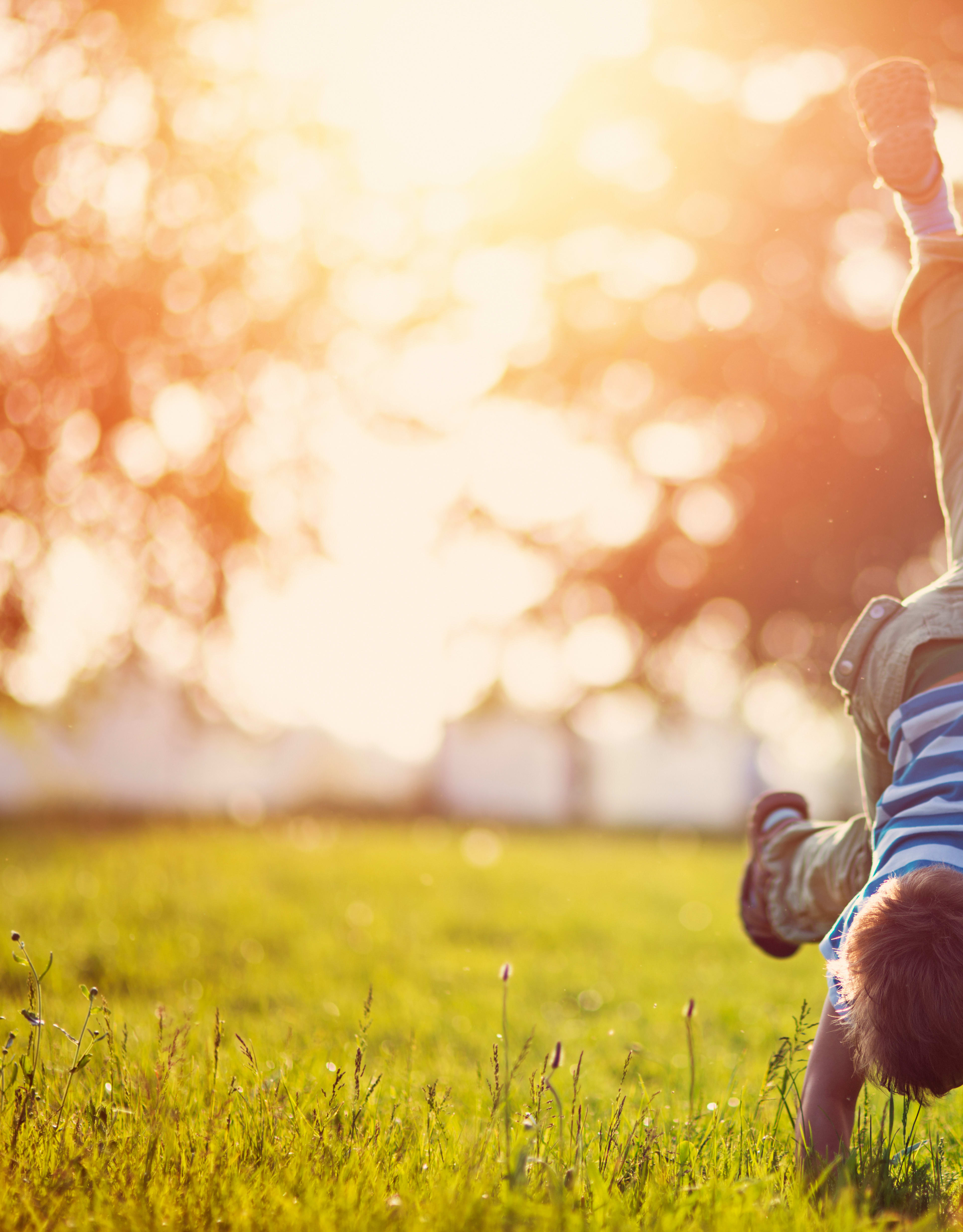 A person in striped clothing is performing a handstand on a grassy field, with a blurred background of trees and sunlight filtering through.