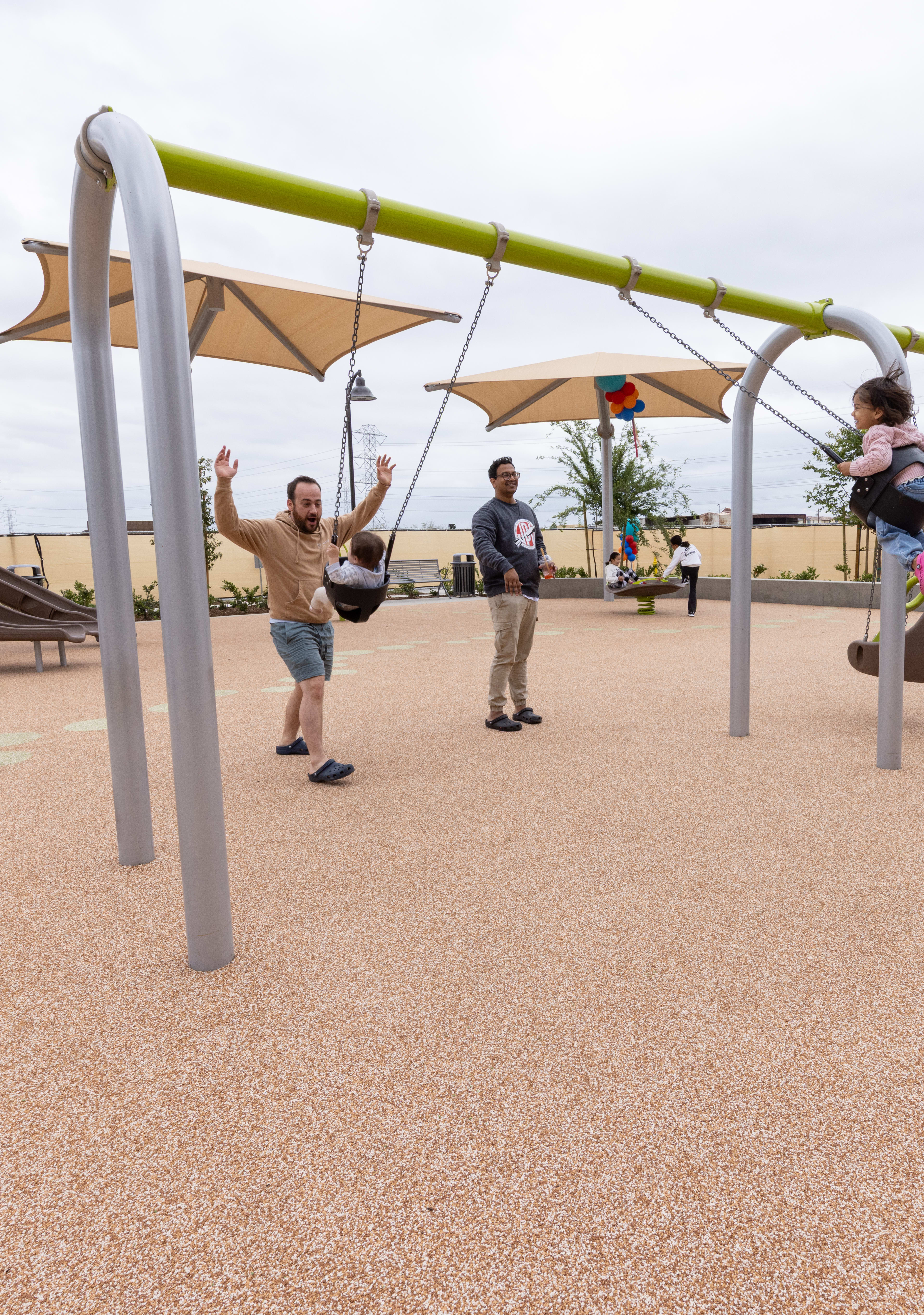 A modern outdoor playground with various colorful equipment and structures, surrounded by residential buildings and shaded areas.