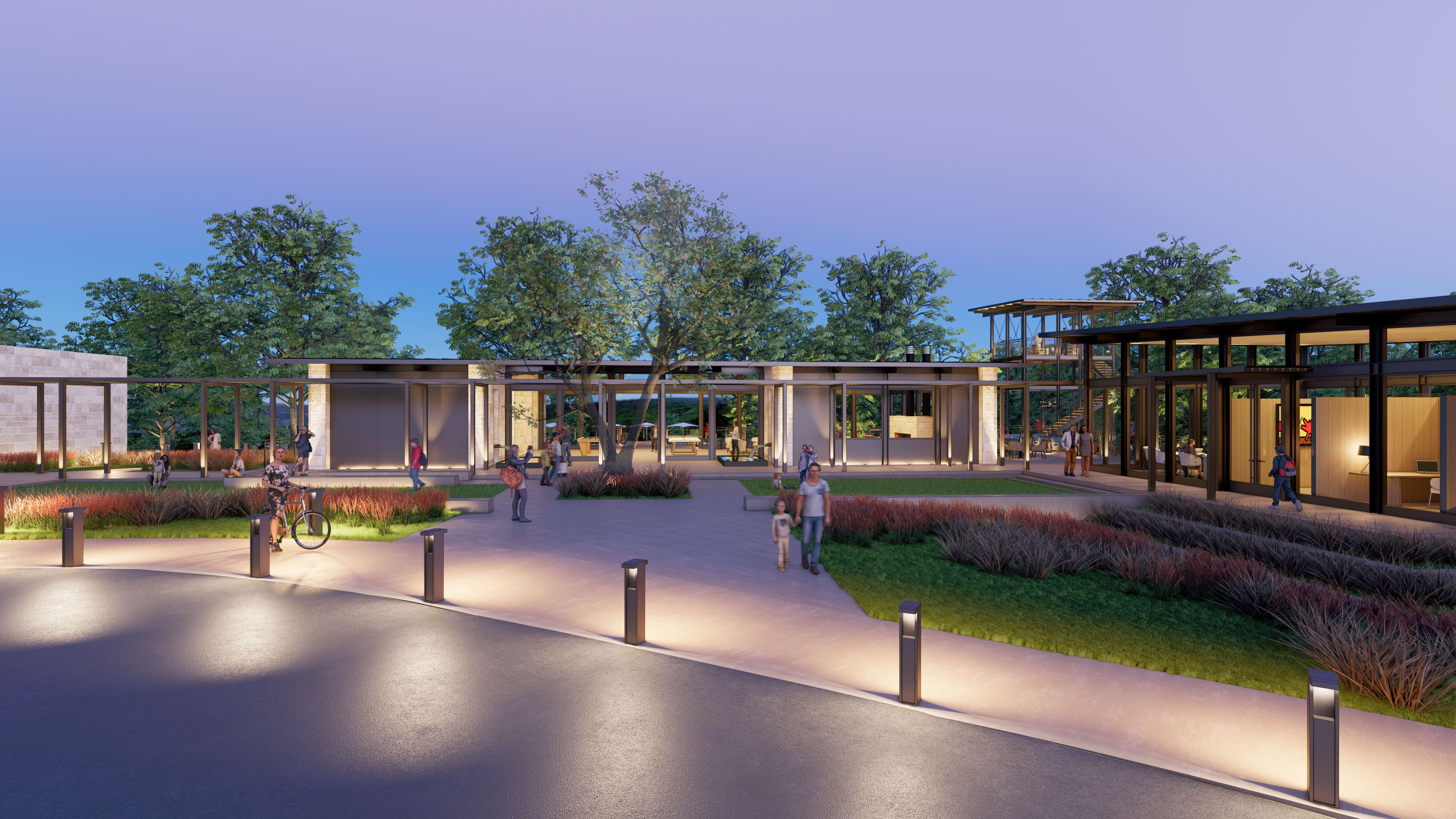 A modern outdoor public space with a paved walkway, landscaped gardens, and several buildings with glass facades surrounded by trees against a twilight sky.