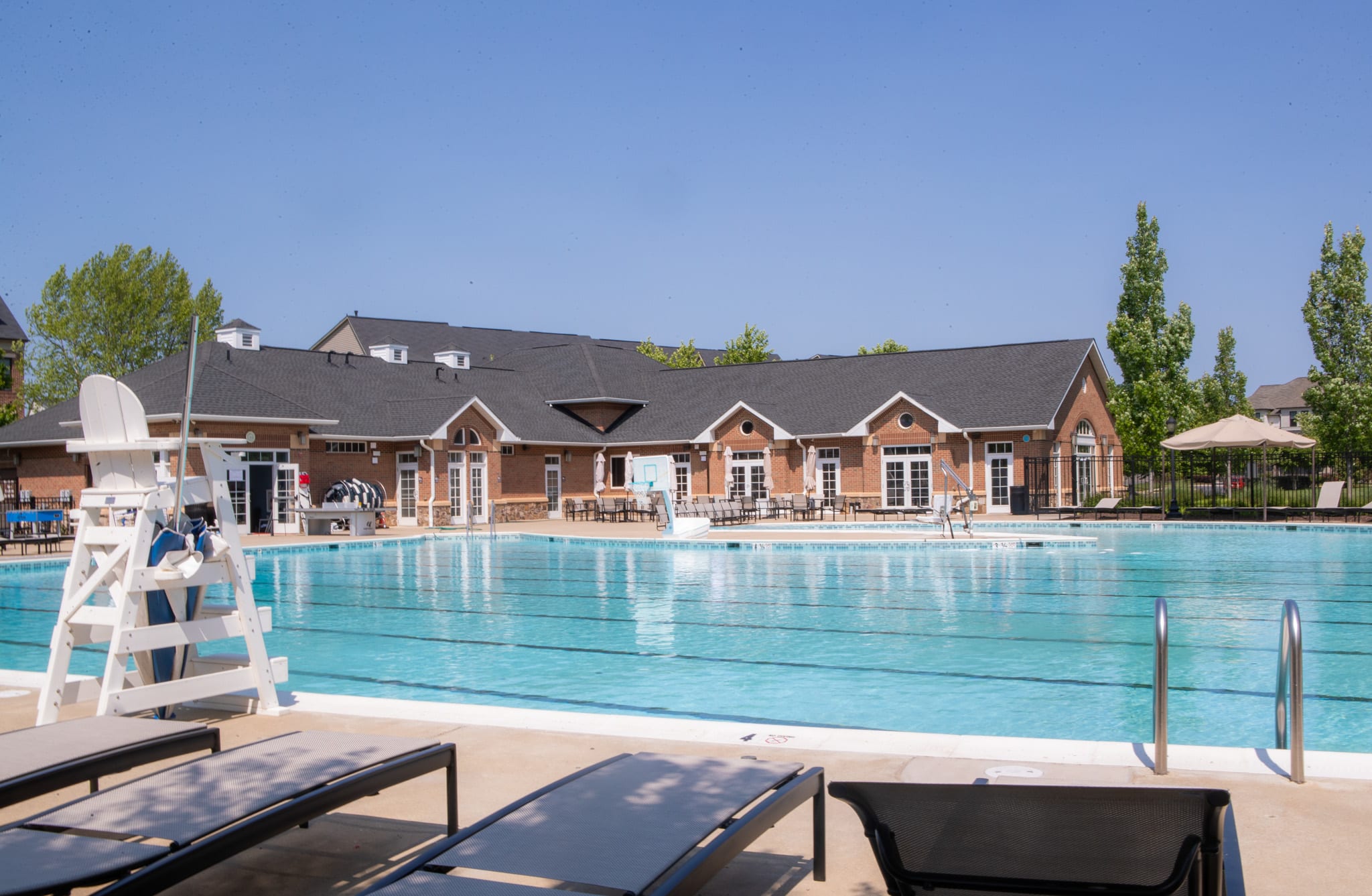 A large outdoor swimming pool surrounded by a brick building with a tiled roof, with lounge chairs in the foreground and a clear blue sky in the background.