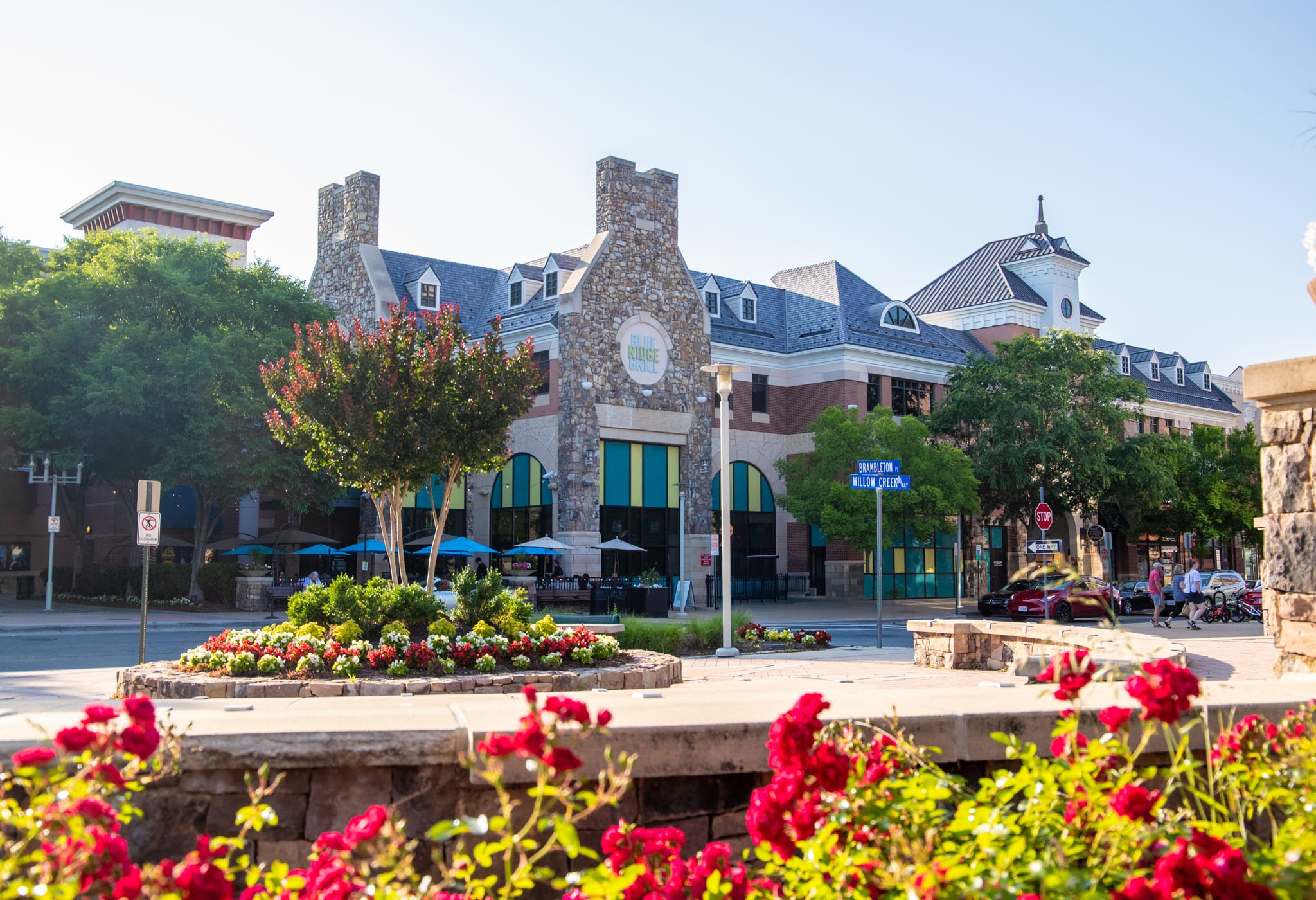 A vibrant and colorful town square with a stone fountain surrounded by blooming flowers, with a mix of historic and modern buildings in the background.