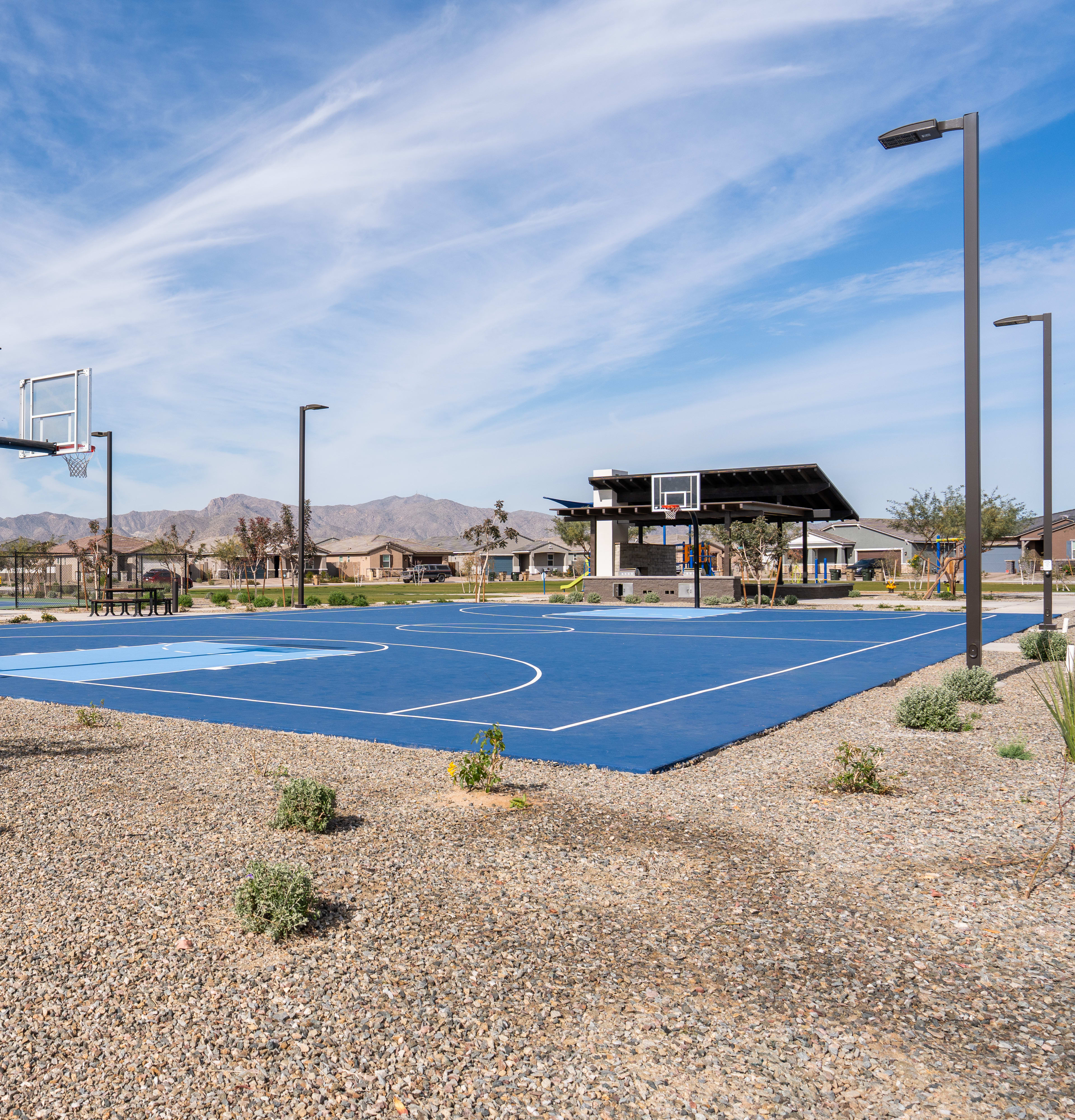 A well-maintained outdoor basketball court with blue and white markings, surrounded by a gravel surface and desert landscape in the background under a clear blue sky with wispy clouds.