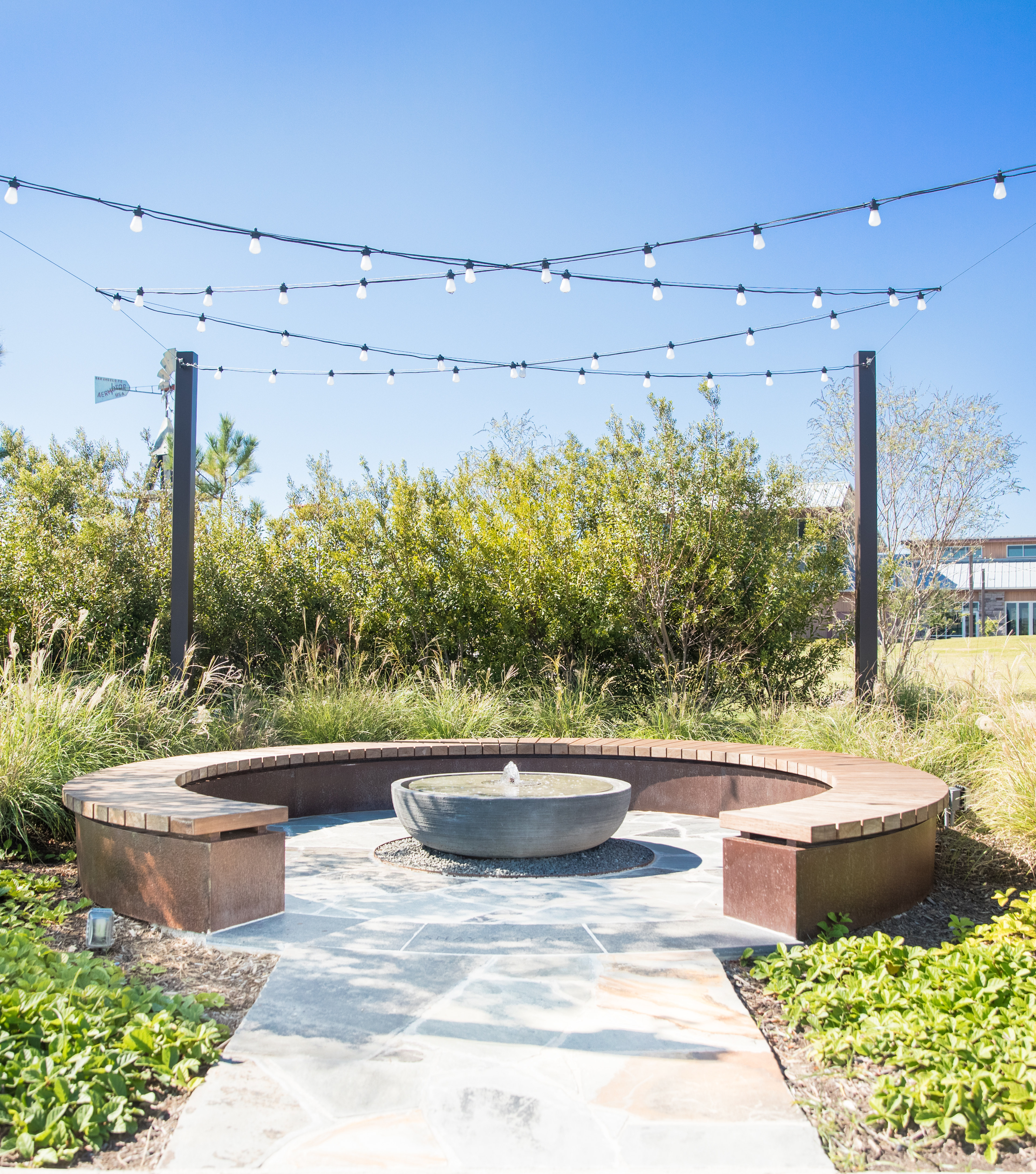 A cozy outdoor seating area with a fire pit surrounded by lush greenery, string lights overhead, and a building visible in the background.