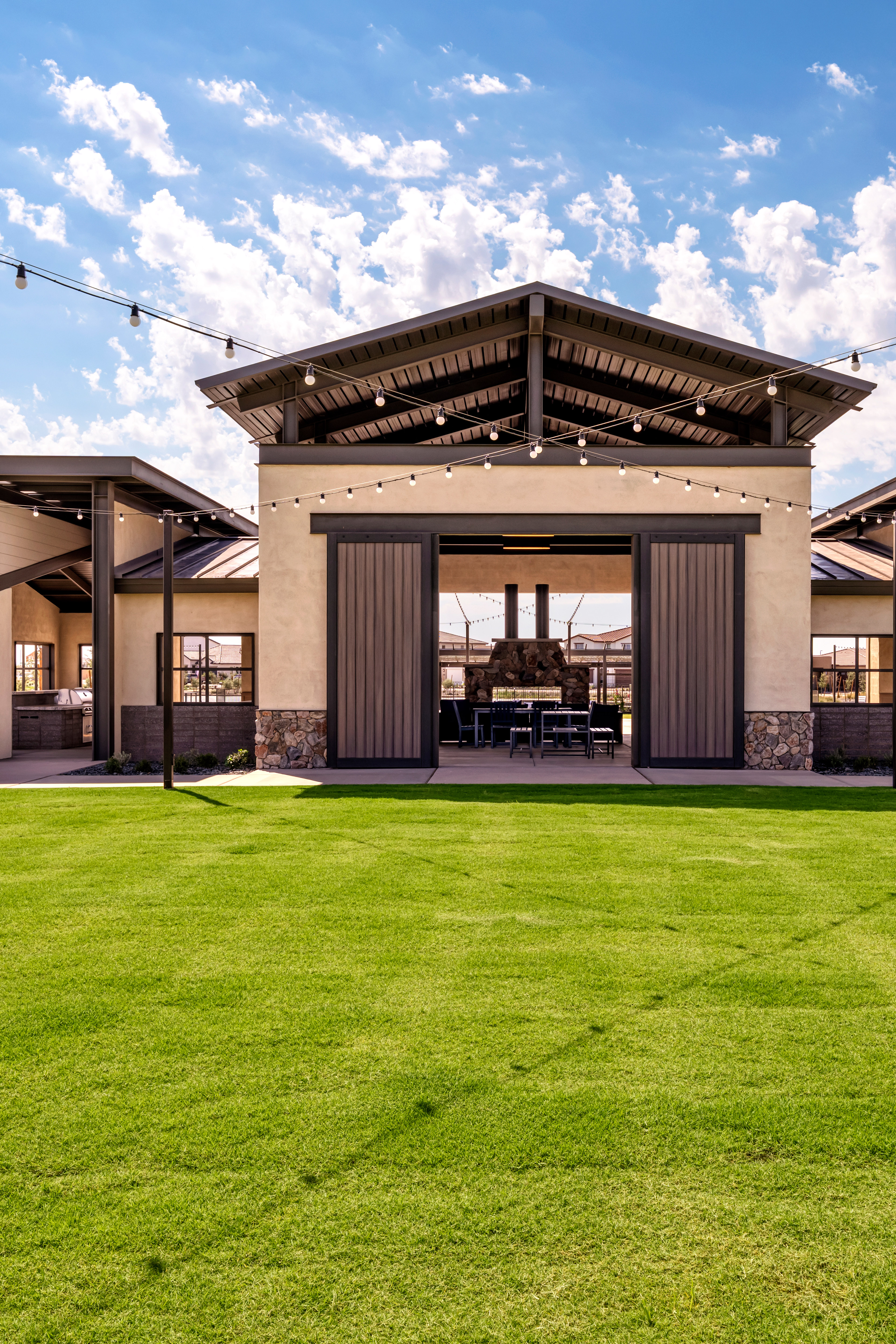 A large, modern building with a wooden exterior stands in the foreground, surrounded by a lush green lawn. The sky in the background is filled with fluffy white clouds against a bright blue backdrop.