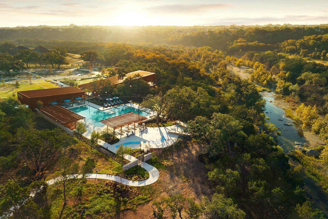 A scenic aerial view of a resort nestled in a lush, forested landscape, with a winding river and swimming pools visible in the foreground, all bathed in the warm glow of the setting sun.