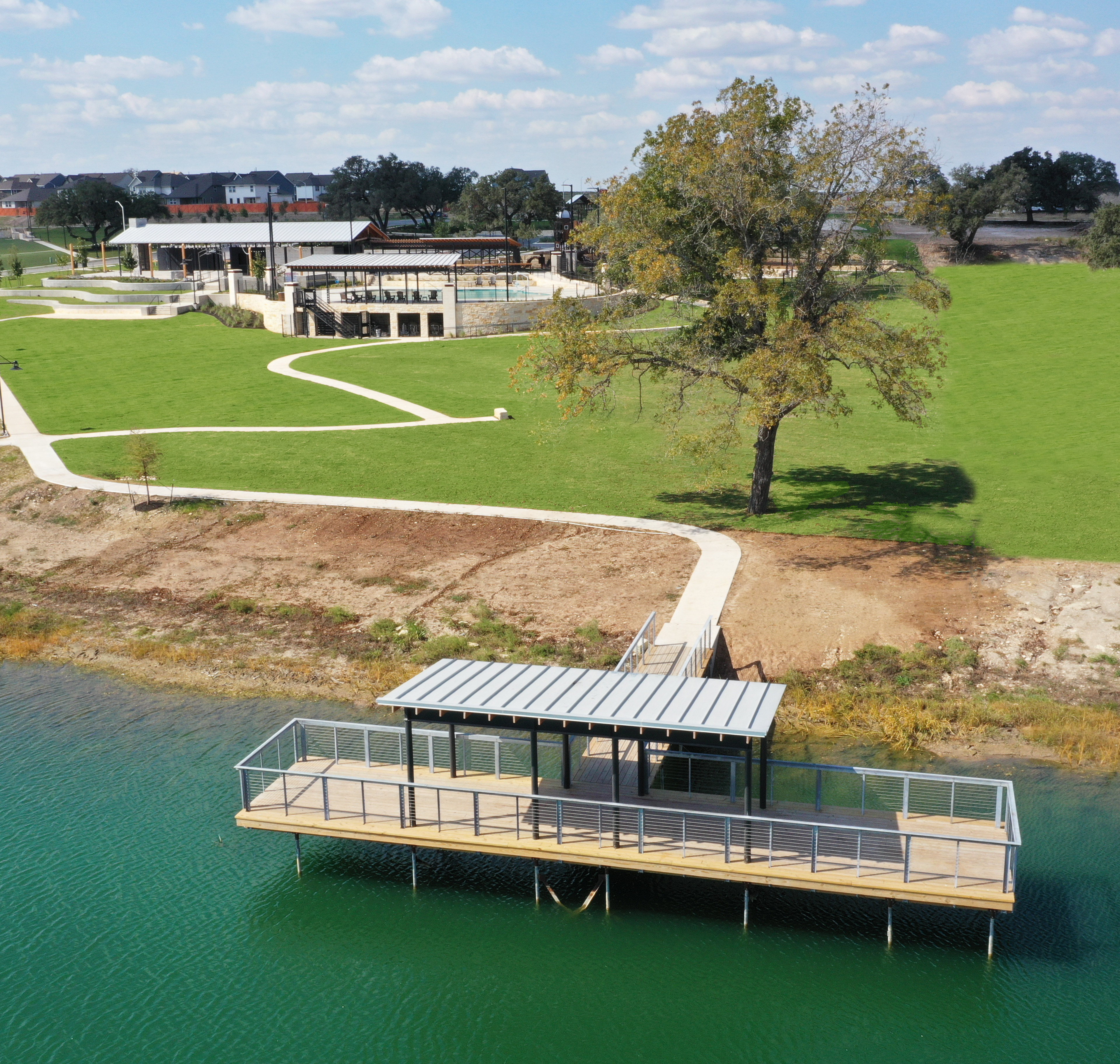 A scenic landscape with a grassy field, a body of water, and a wooden dock structure in the foreground, surrounded by trees and buildings in the background.