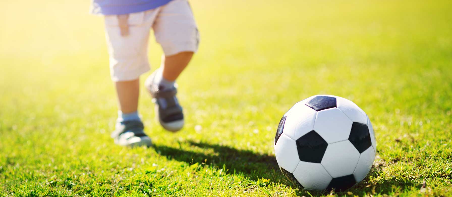 A soccer ball rests on a lush, grassy field as a person in athletic attire approaches it, suggesting a scene of an upcoming soccer game or practice.