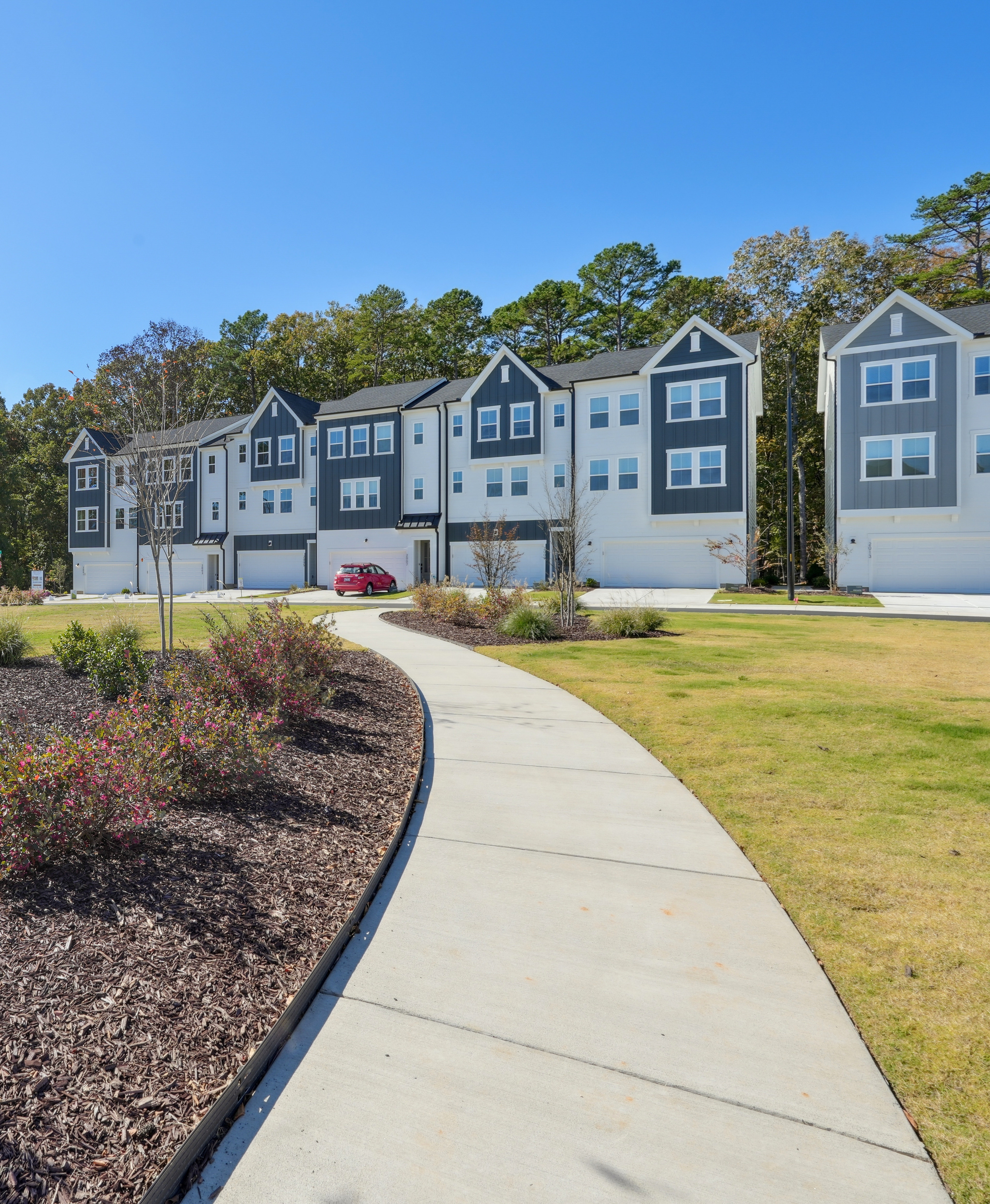 A paved walkway leads through a well-manicured landscape of shrubs and trees, with a row of multi-story townhouses visible in the background against a clear blue sky.