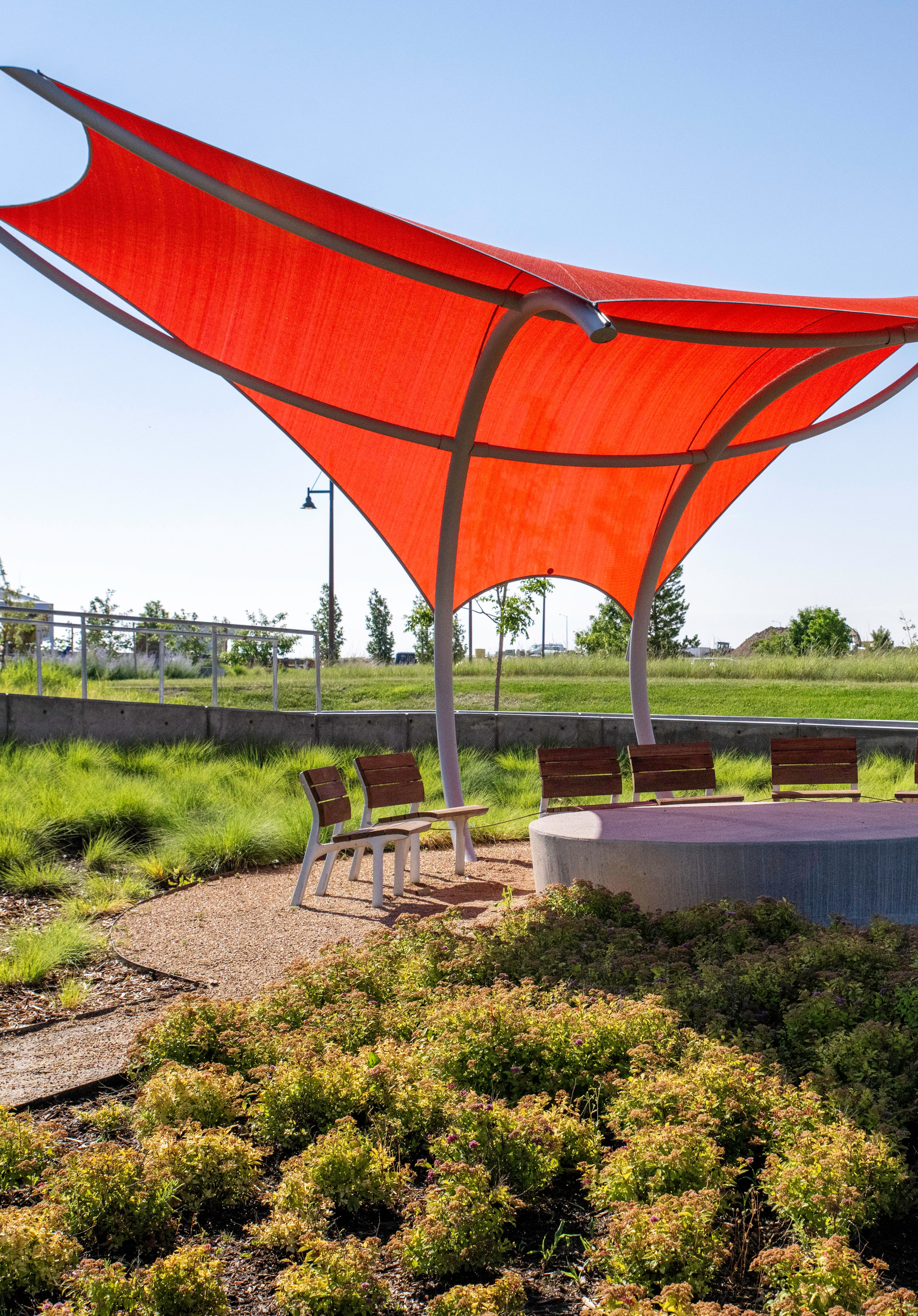 A large, vibrant red canopy structure stands in a landscaped outdoor area, surrounded by lush greenery and a paved seating area.