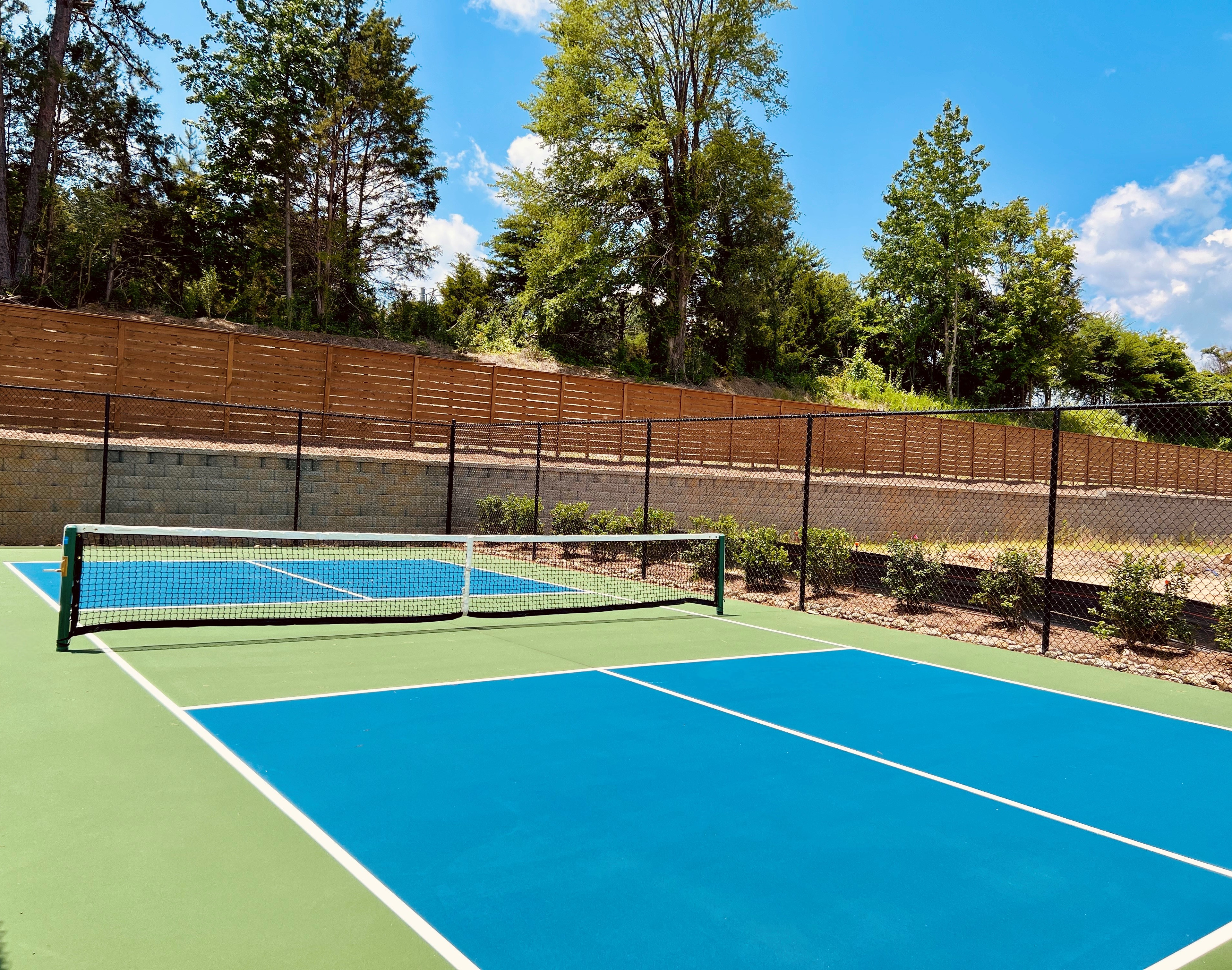 A tennis court with a blue and green surface surrounded by a wooden fence, with a forested area in the background.