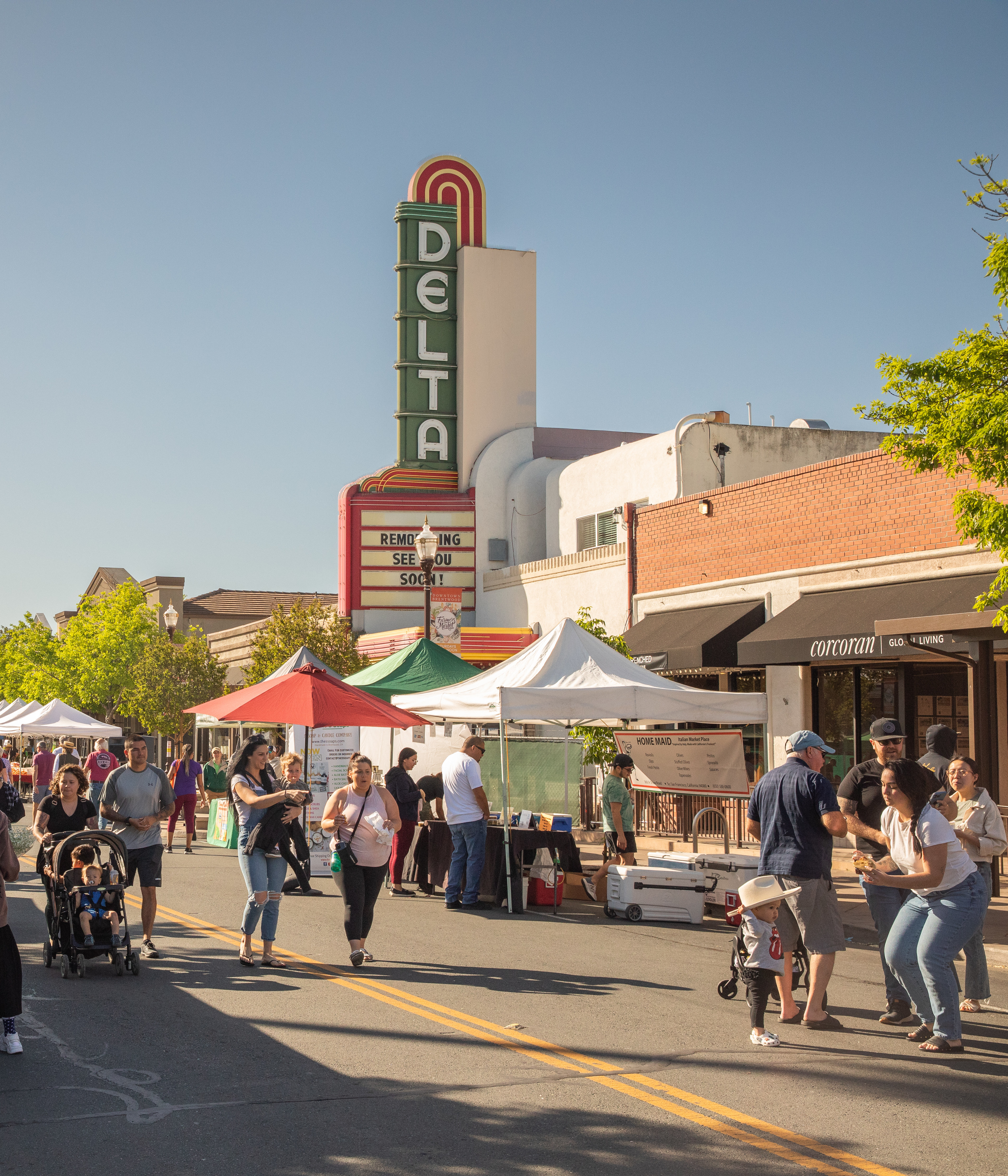 A bustling outdoor market with various vendors and tents, set against the backdrop of a historic Delta theater building with its iconic neon sign.