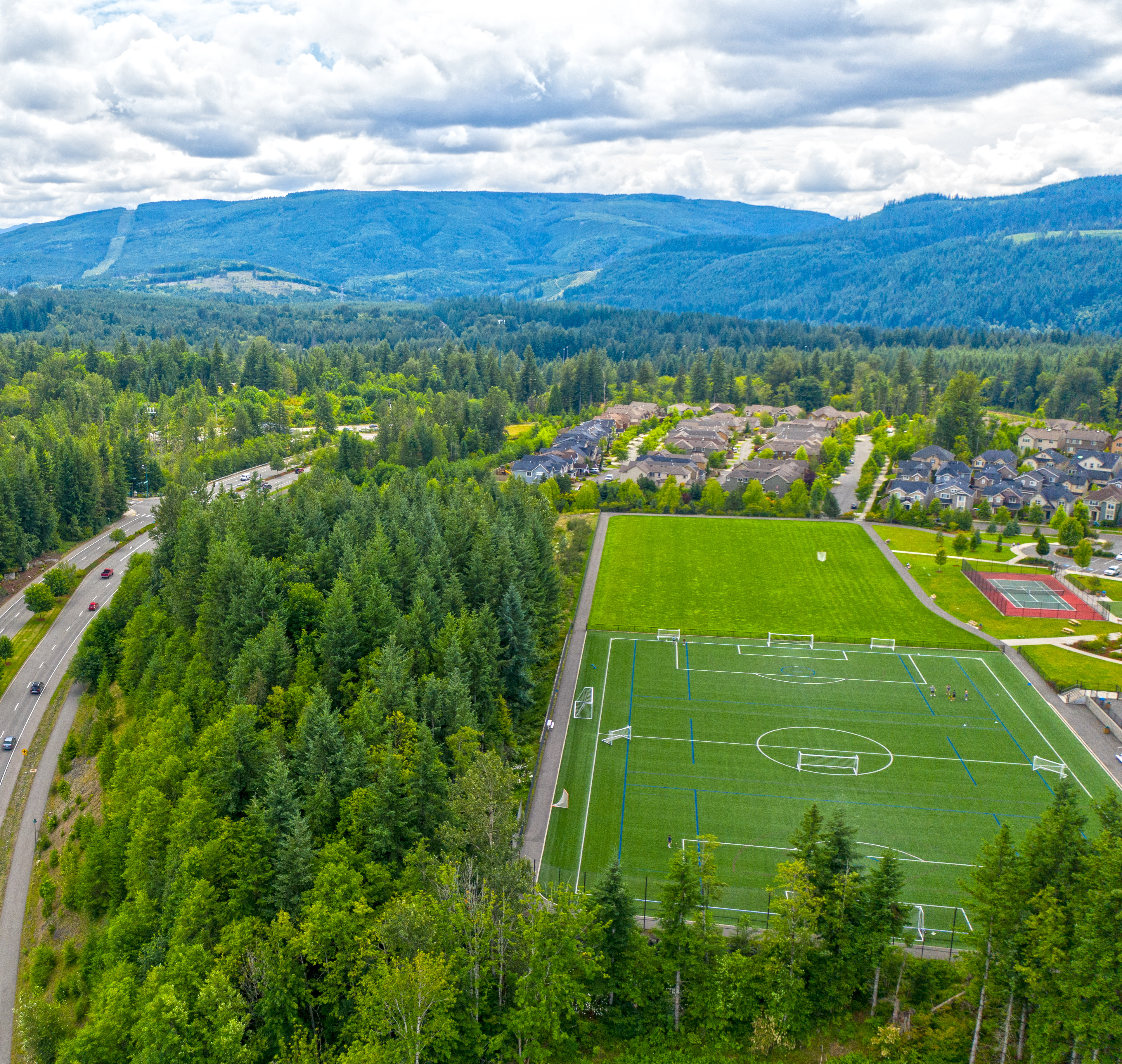A lush, forested landscape with a soccer field in the foreground, surrounded by winding roads and towering mountains in the background under a cloudy sky.