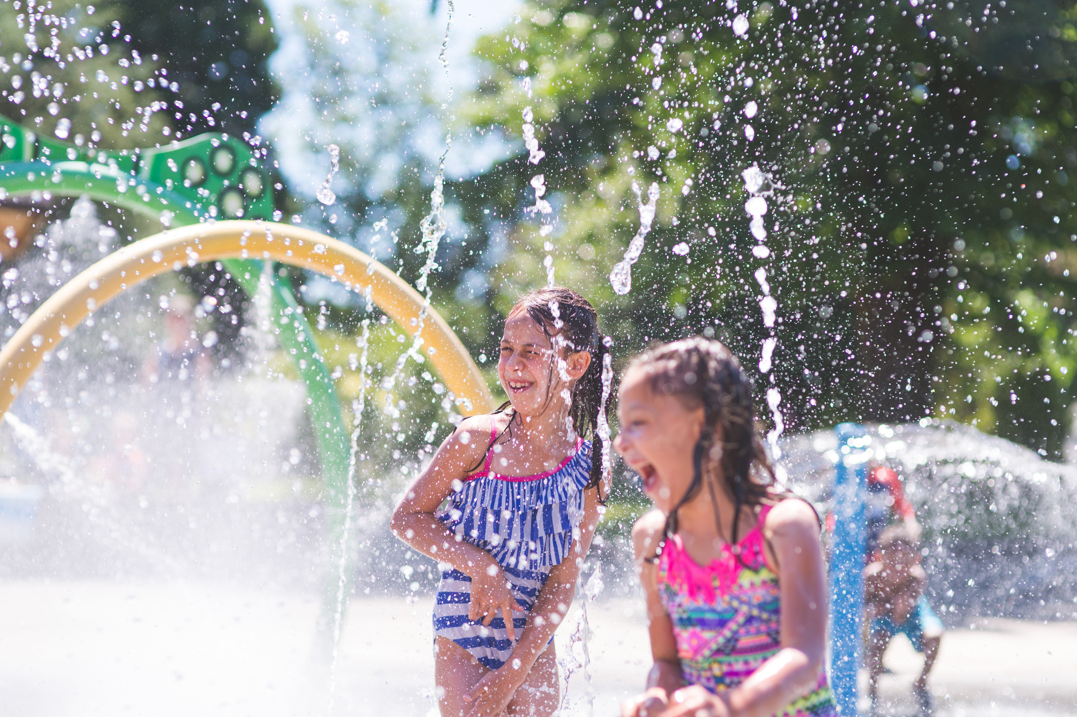 Two young girls playing in a colorful water fountain, surrounded by lush greenery and a vibrant, playful atmosphere.