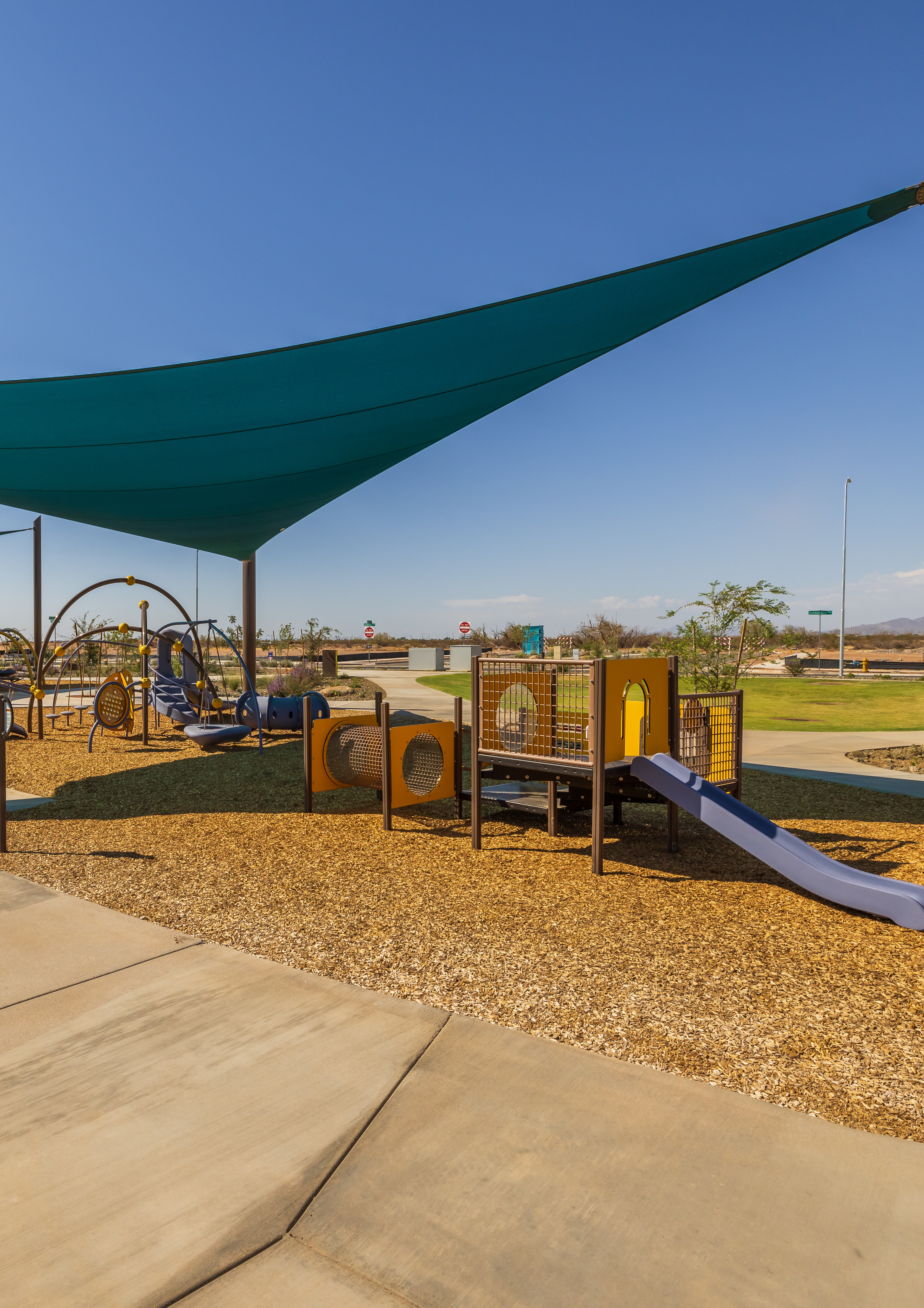 A shaded playground area with various play equipment, surrounded by a grassy landscape and a clear blue sky.