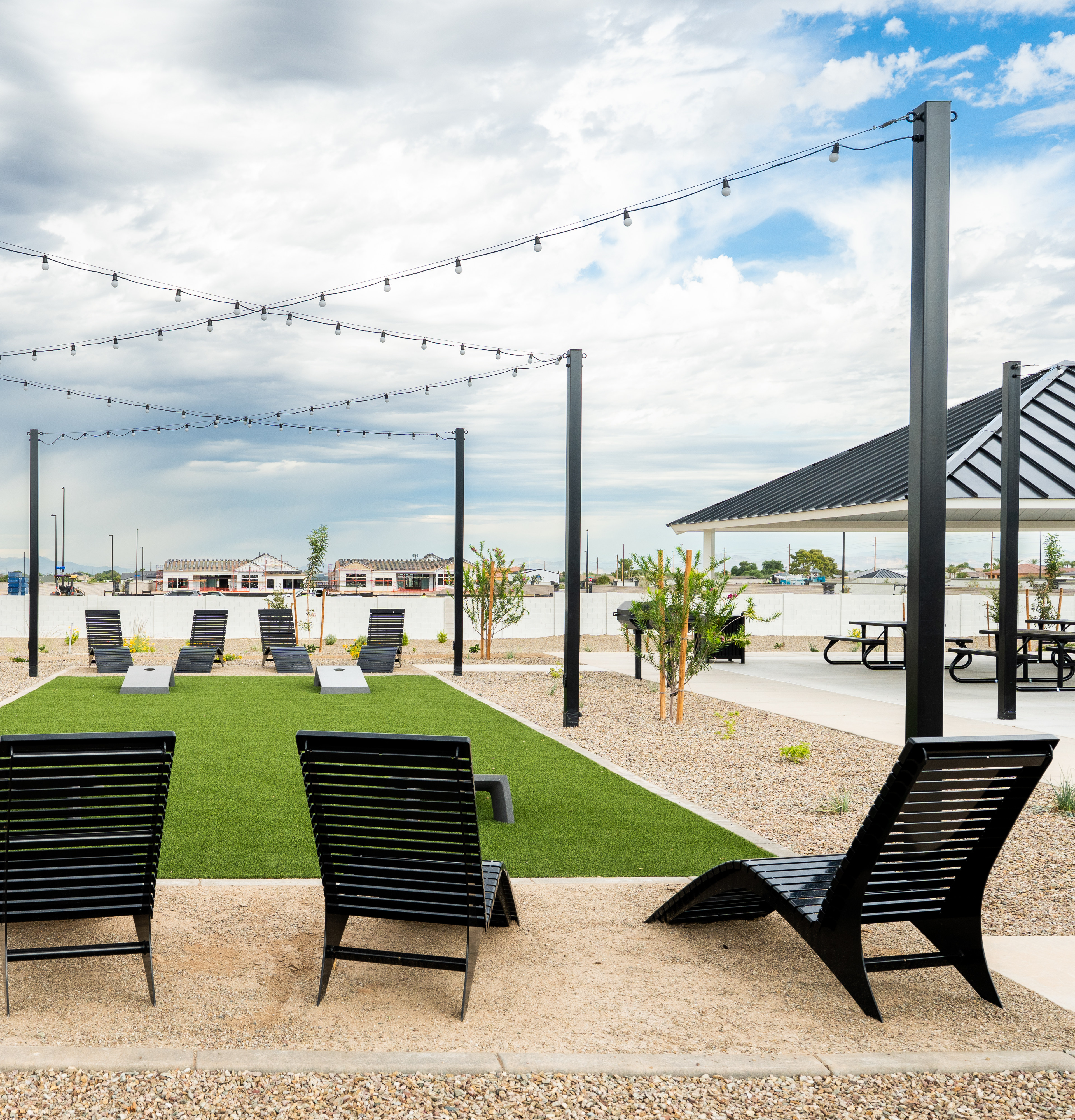 An outdoor seating area with black lounge chairs arranged on a grassy surface, surrounded by metal frames supporting string lights and a cloudy sky in the background.