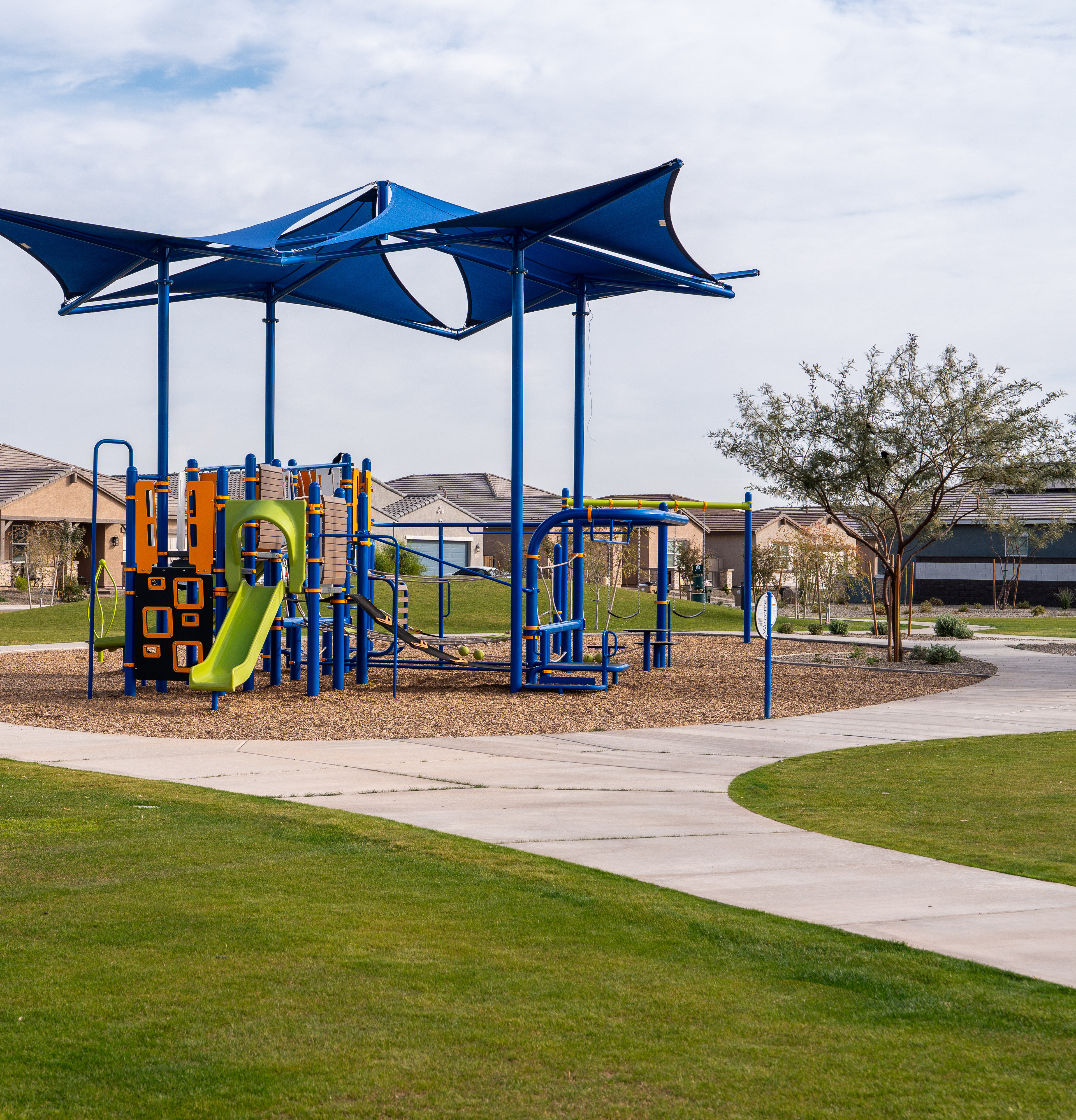 A colorful playground with various play structures, including slides and climbing equipment, surrounded by a grassy area and residential buildings in the background.